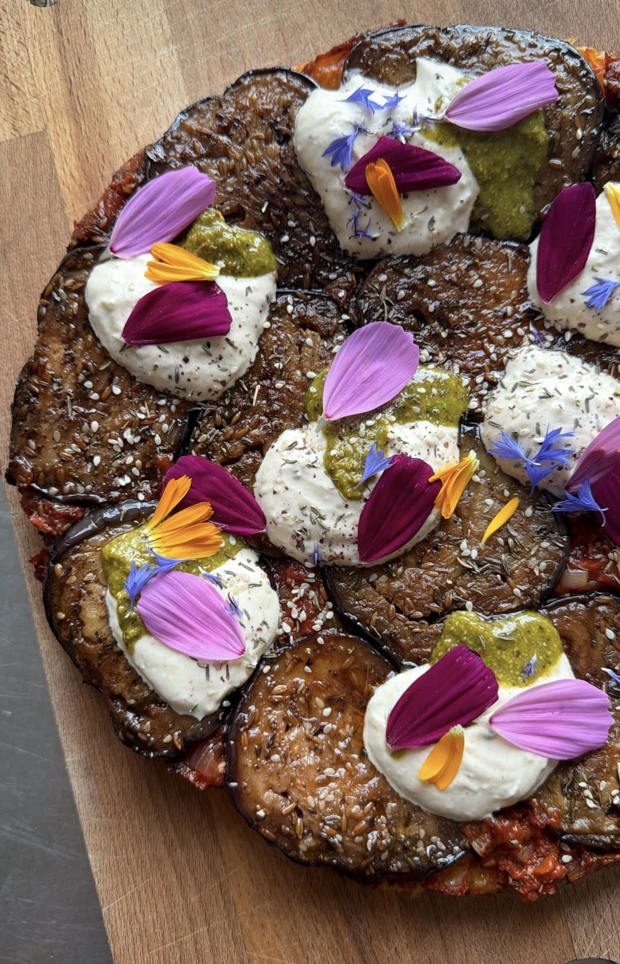 A vegetable pizza topped with dollops of white cheese, edible flower petals in pink, purple, orange, and yellow, green pesto, and sesame seeds, on a wooden surface.
