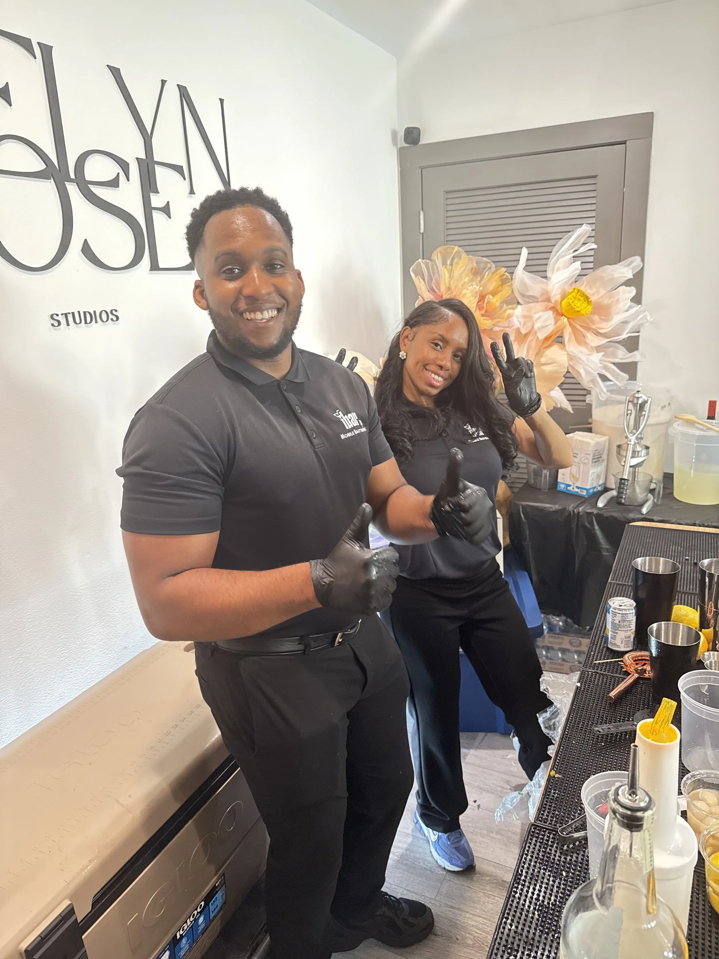 Two people wearing black shirts and gloves are smiling and making peace signs behind a counter with drink bottles and utensils, in a room with a decorative floral background and the words 'STUDIOS' on the wall.