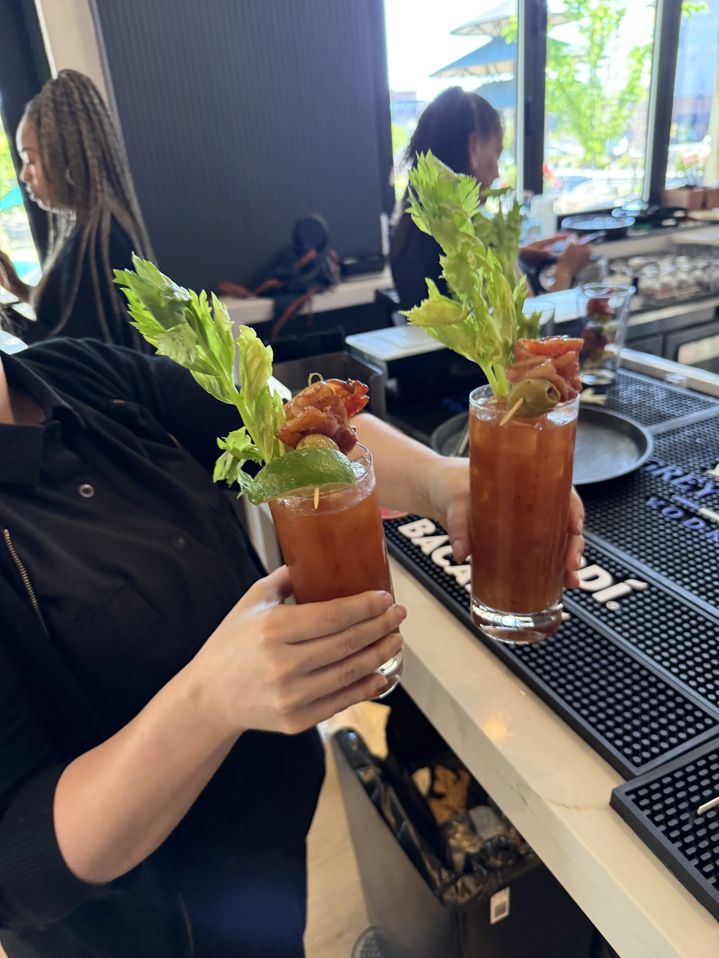 Two hands holding up margarita glasses topped with celery, bacon, and lime, inside a restaurant or bar with large windows and a view outside.