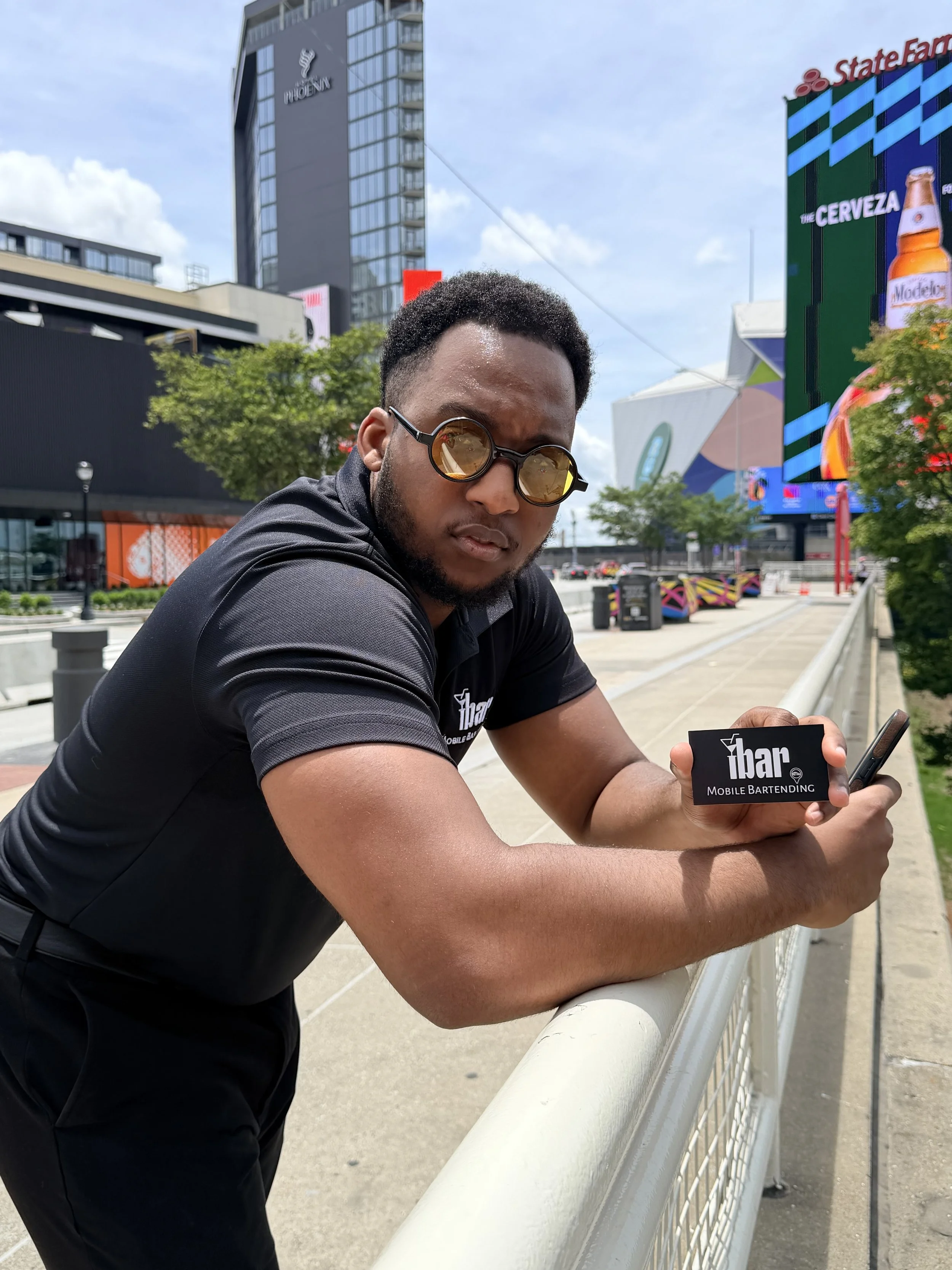 A man wearing sunglasses and a black shirt, leaning on a white railing outdoors in an urban area, holding a business card for 'iBar Mobile Bartending'.