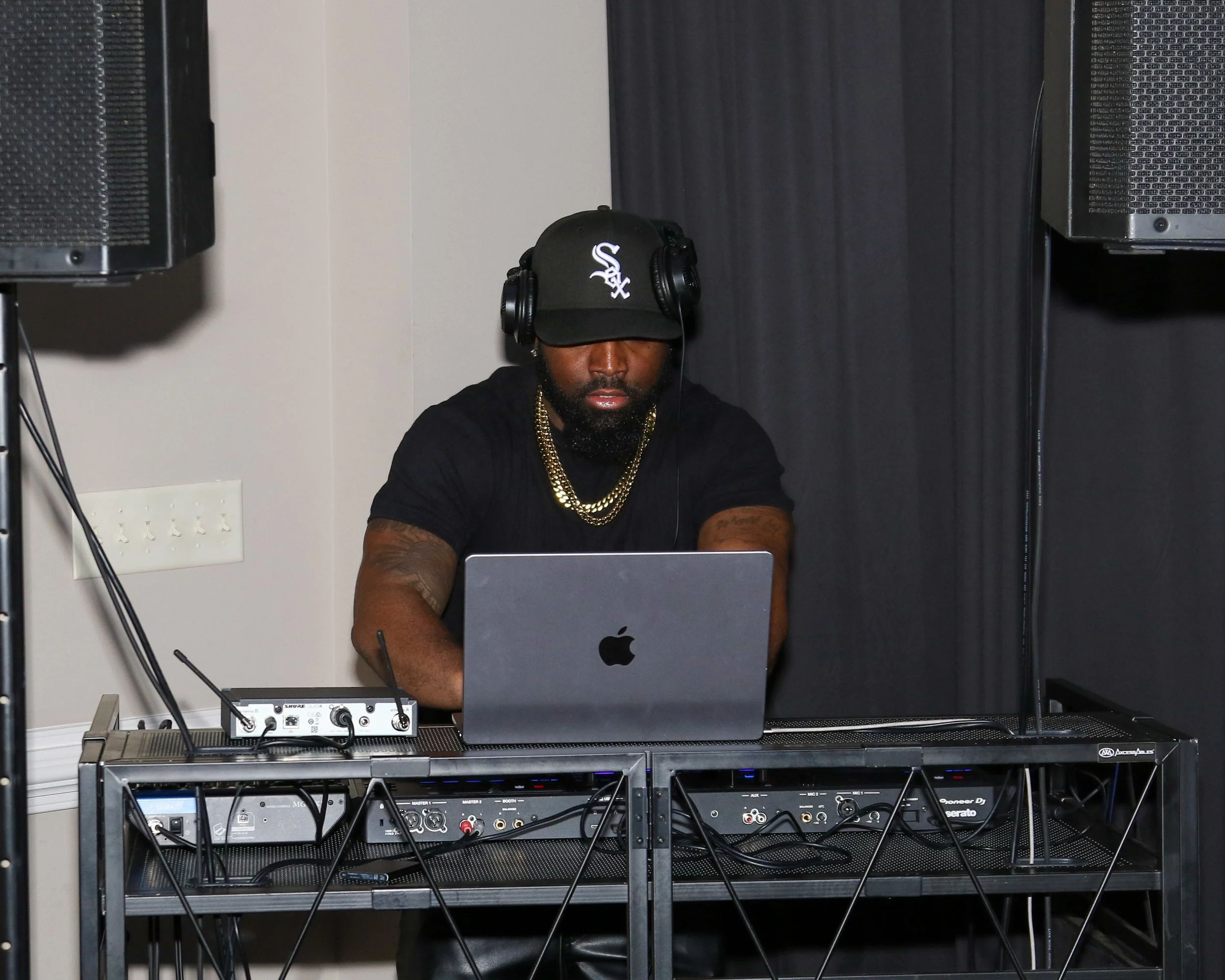 A man wearing a black baseball cap with a white logo, headphones, and gold chains around his neck is working on a music production setup. He is focused on a laptop in front of him, with audio equipment and speakers visible on a stand and a rack below