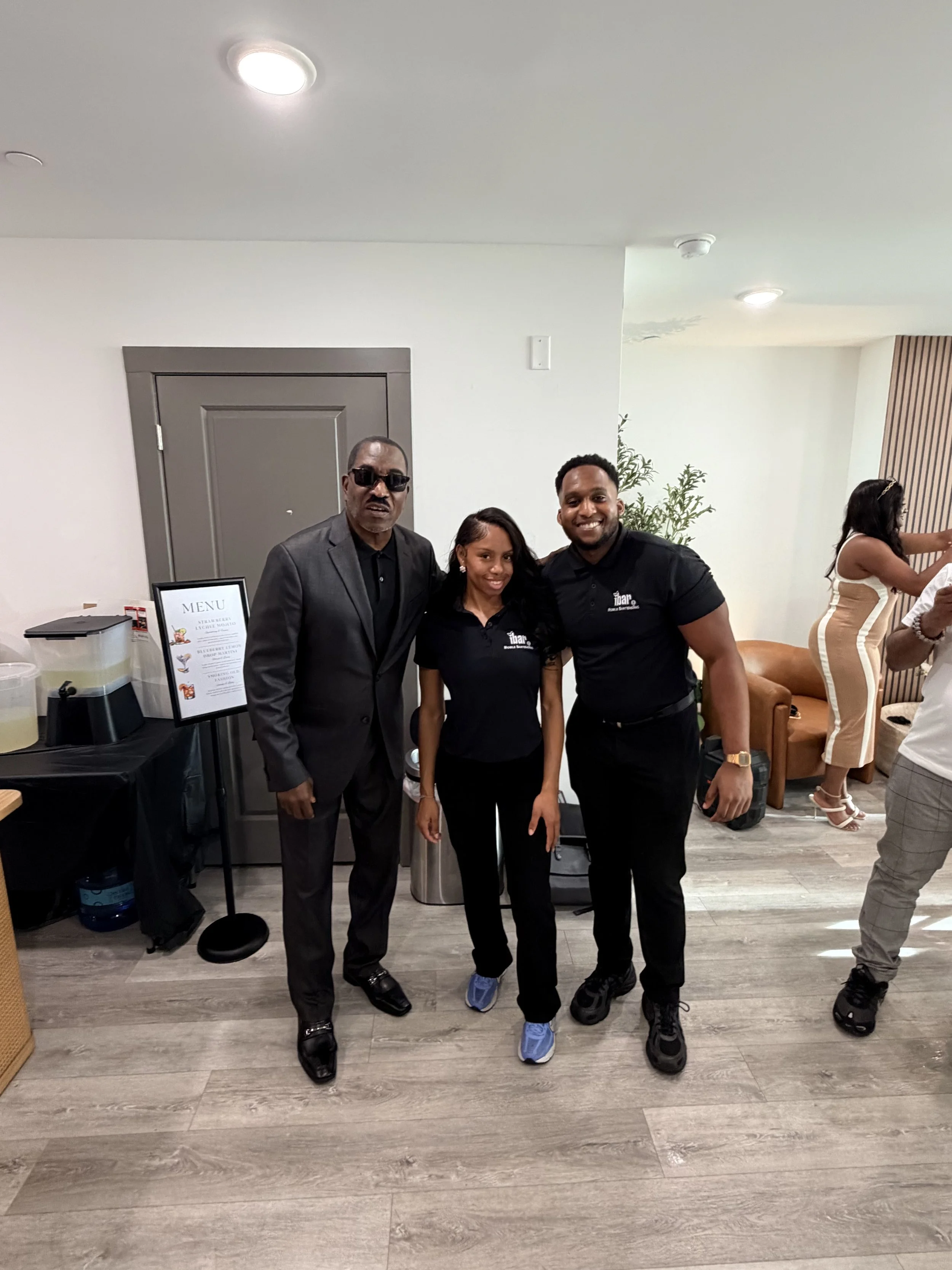 Three people standing together indoors, posing for a photo. The man on the left is wearing sunglasses and a dark suit, the woman in the middle is smiling and dressed in black, and the man on the right is smiling and wearing a black polo shirt with a 