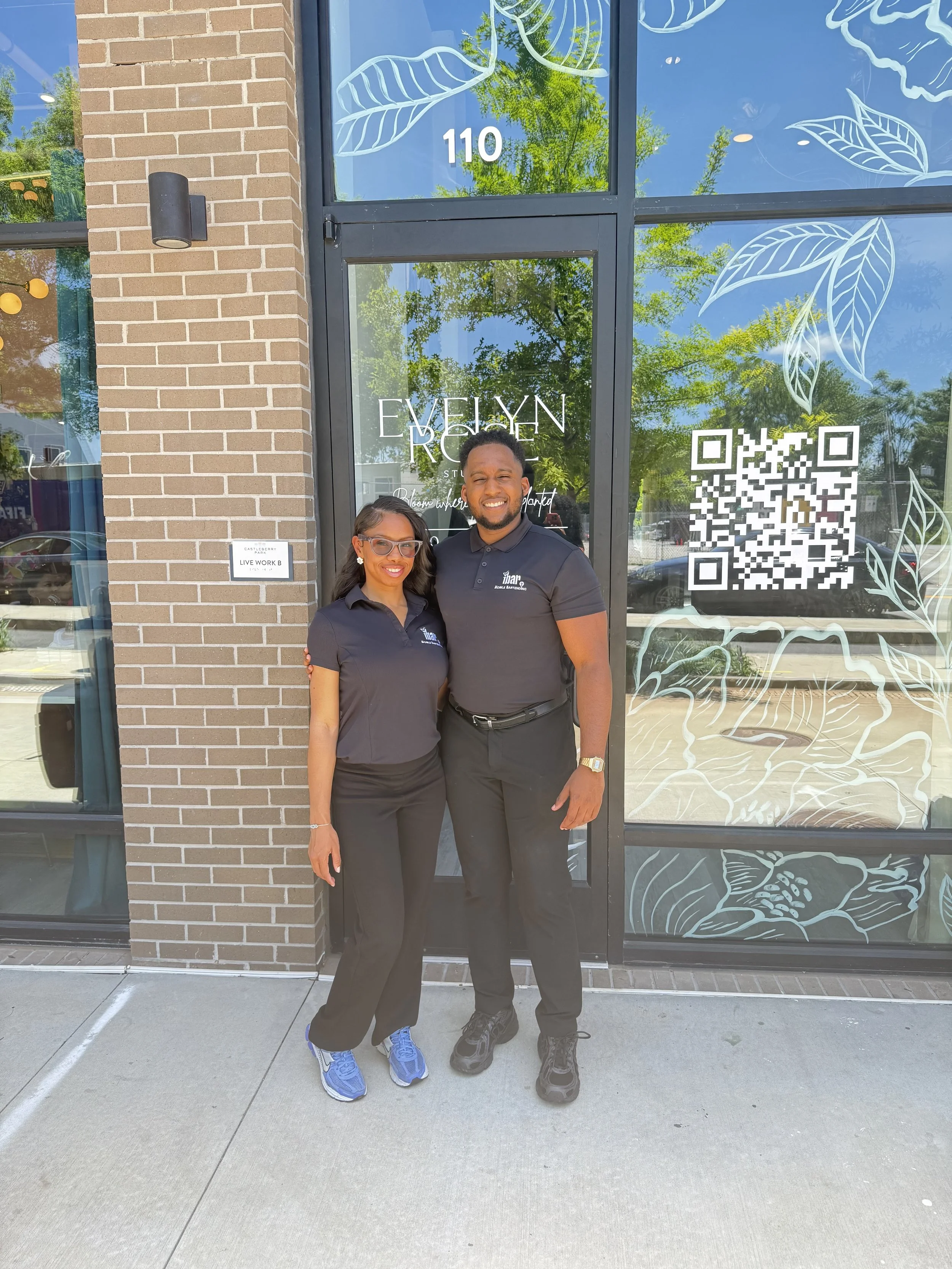 Two smiling people standing outside in front of a store window, which has a leaf design and the address 110 on it. The woman on the left is wearing glasses, a dark polo shirt, black pants, and blue sneakers. The man on the right is wearing a black po