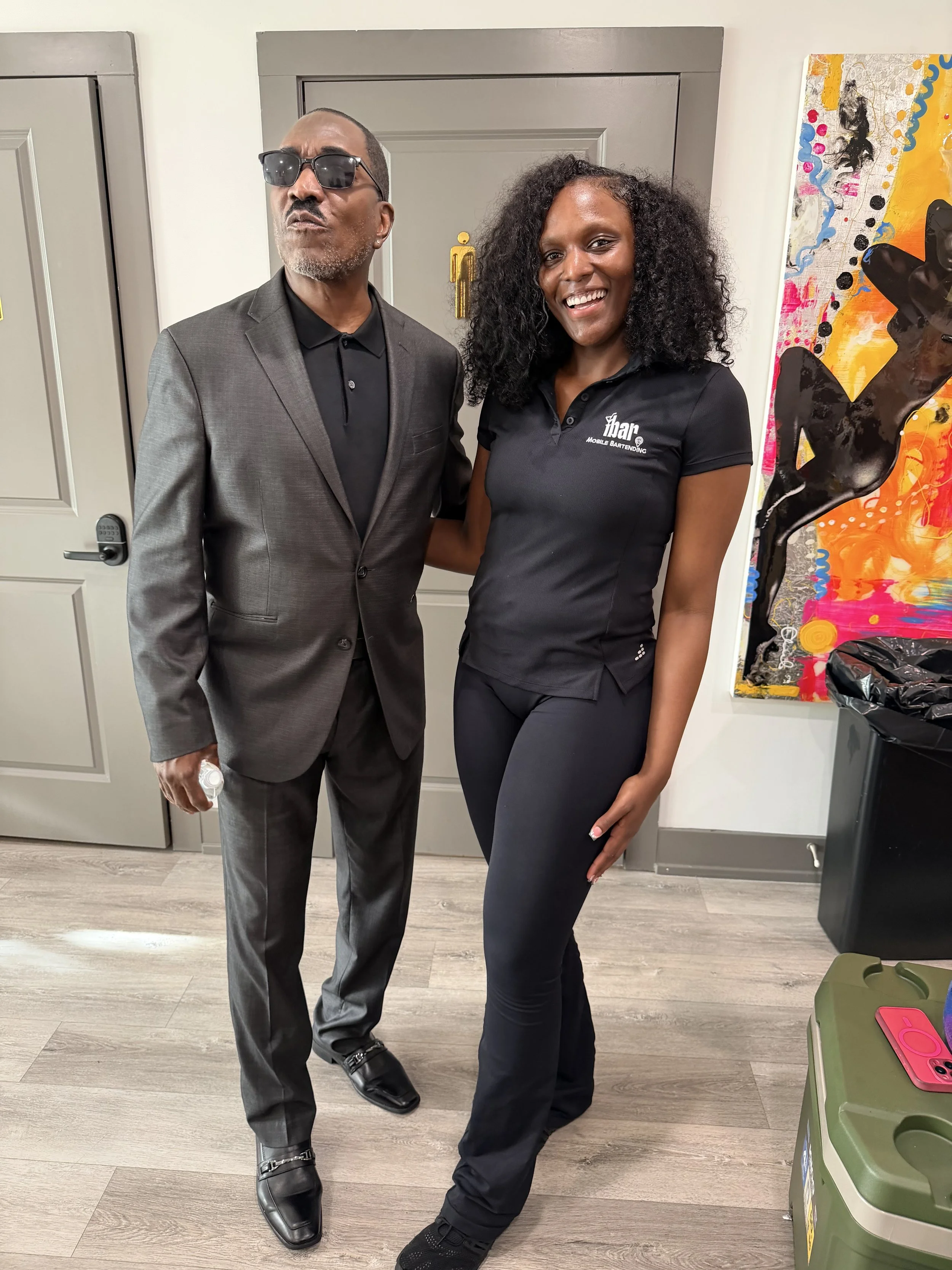 A man in a gray suit with sunglasses and a woman in a black polo shirt and black pants standing together indoors, smiling at the camera, with colorful abstract art on the wall behind them.