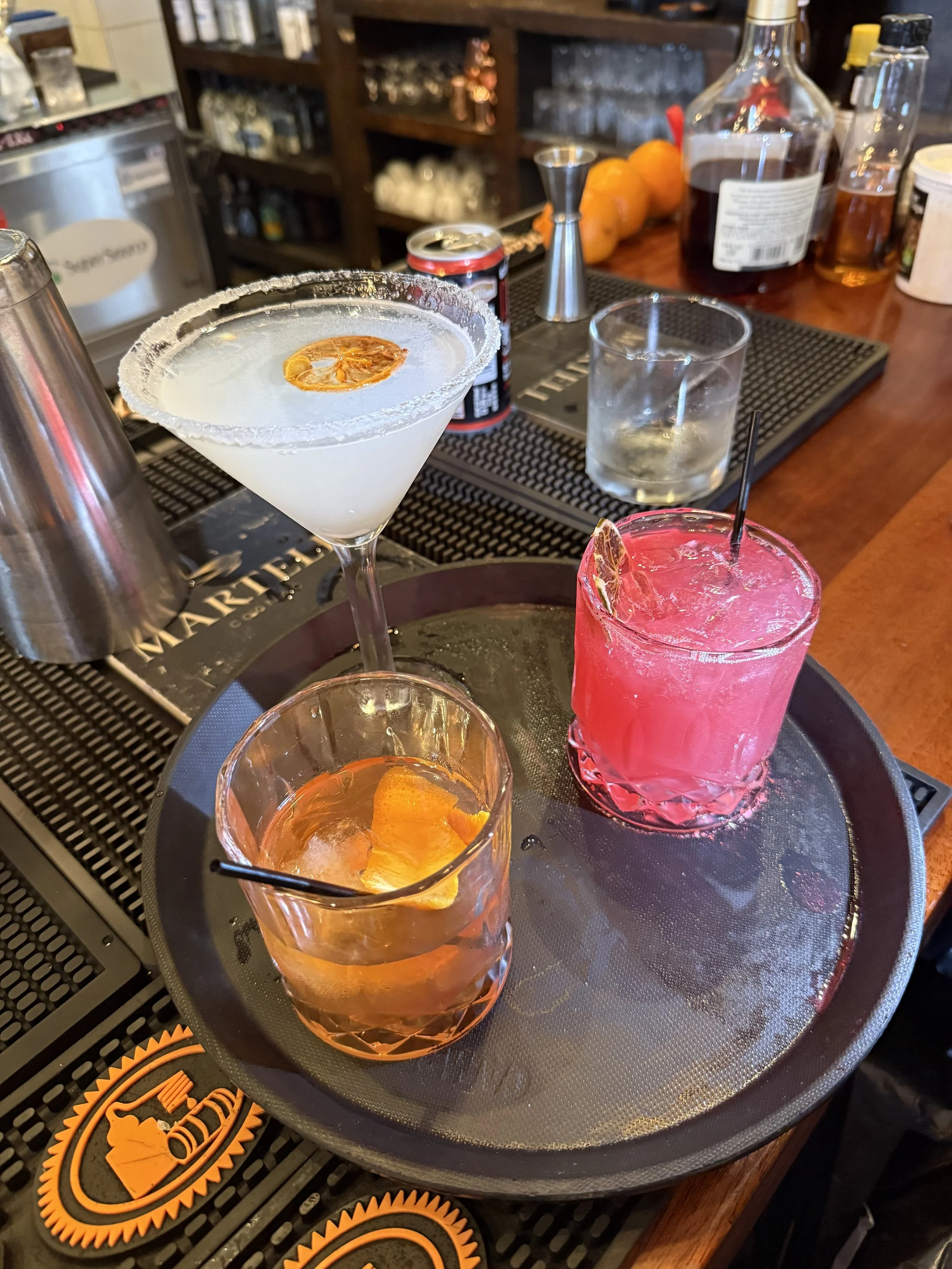Photograph of a bar counter with various colorful cocktails, including a white cocktail with a salted rim and a citrus garnish, a pink cocktail, and a glass with a citrus slice. The background shows bar shelves with bottles and a jigger.