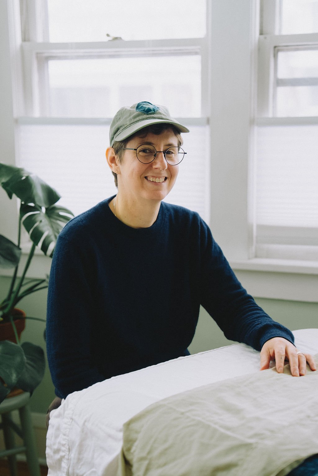 A person with short hair, wearing glasses, a green cap, and a navy blue sweater, sitting at a white table in front of a window. They are smiling and resting their right hand on the table, with a green plant visible in the background.
