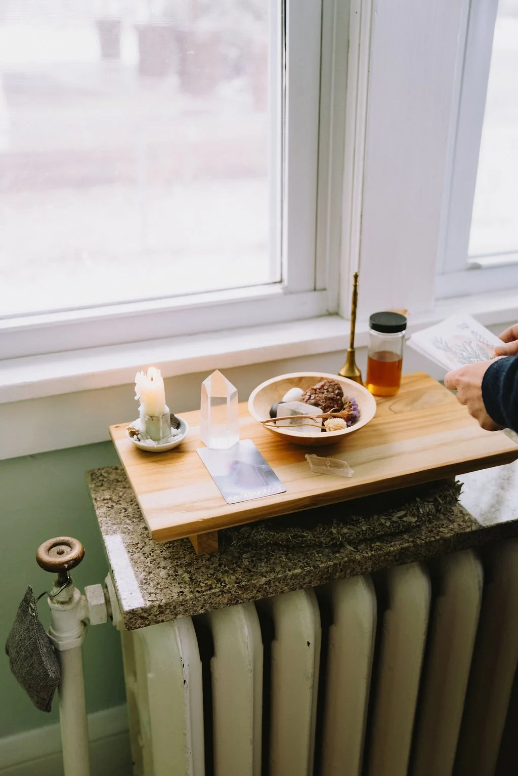 A small wooden table on top of a granite radiator cover with a lit candle, a clear crystal, a bowl with herbs, a glass jar with liquid, and a person holding a paper near a large window.