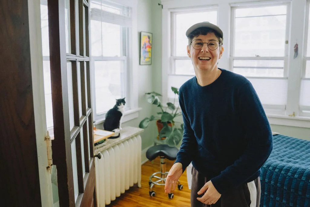 A woman with short hair, glasses, and a cap smiling and laughing in a bright room with a bed, window, and a cat sitting on the windowsill.