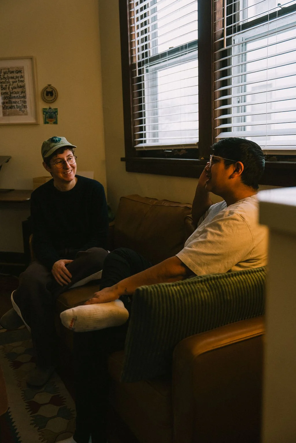 Two people sitting on a couch having a conversation in a cozy room, with windows and blinds behind them.