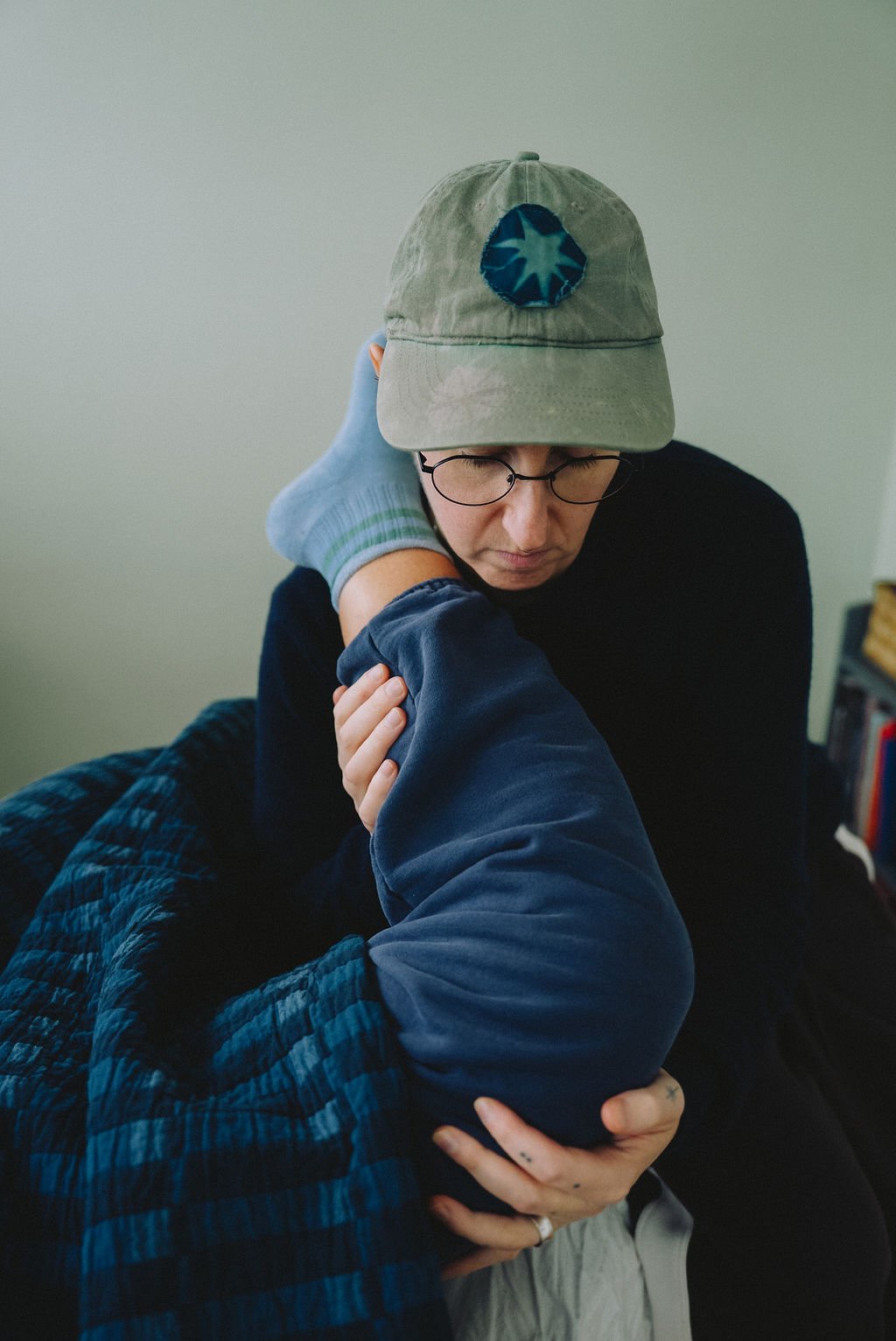A person with glasses, wearing a cap and black sweater, is leaning forward while another person, wearing a blue plaid shirt and sock, is stretching their leg, with the foot near the person's head.