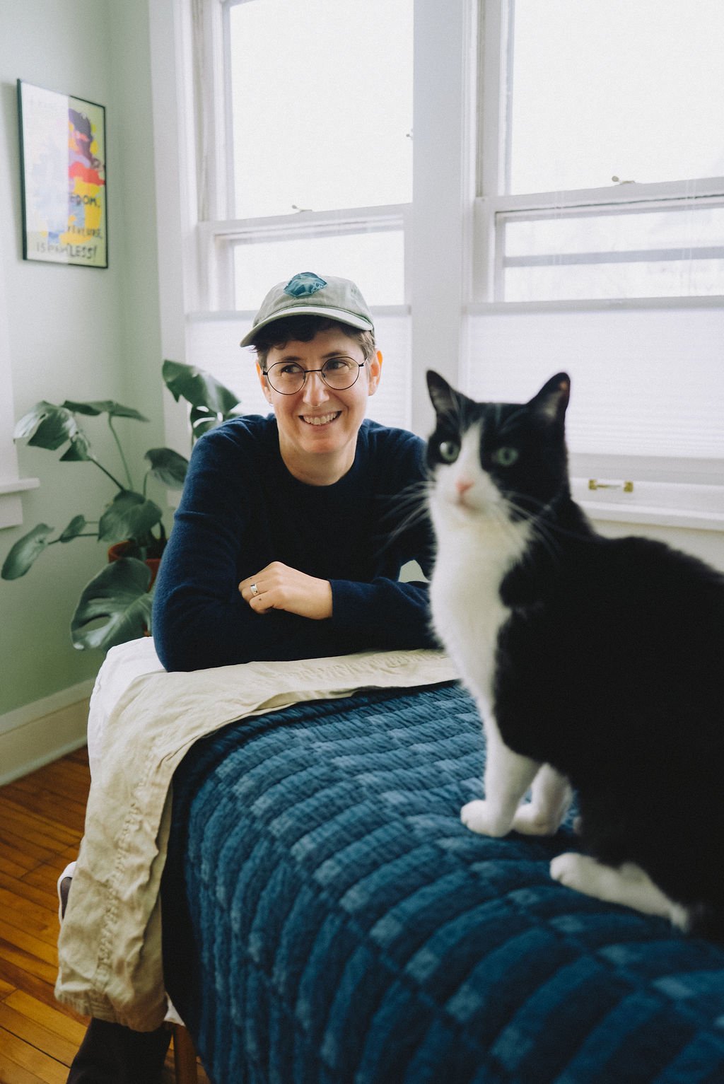 A woman with short dark hair, glasses, and a gray cap is smiling while leaning on a bed. A black and white cat is sitting on the bed in front of her. The room has light green walls, large windows, a plant, and a colorful framed poster on the wall.
