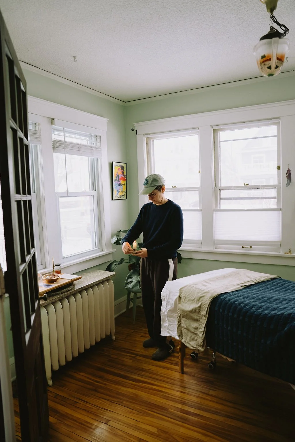 A person in a dark sweater and sweatpants, wearing a gray cap, standing in a room with light green walls and hardwood floors. They are looking down, holding an object. The room has three large windows, a bed with white and dark blue bedding, a green chair, a small table with items, a radiator, and a framed colorful poster on the wall.