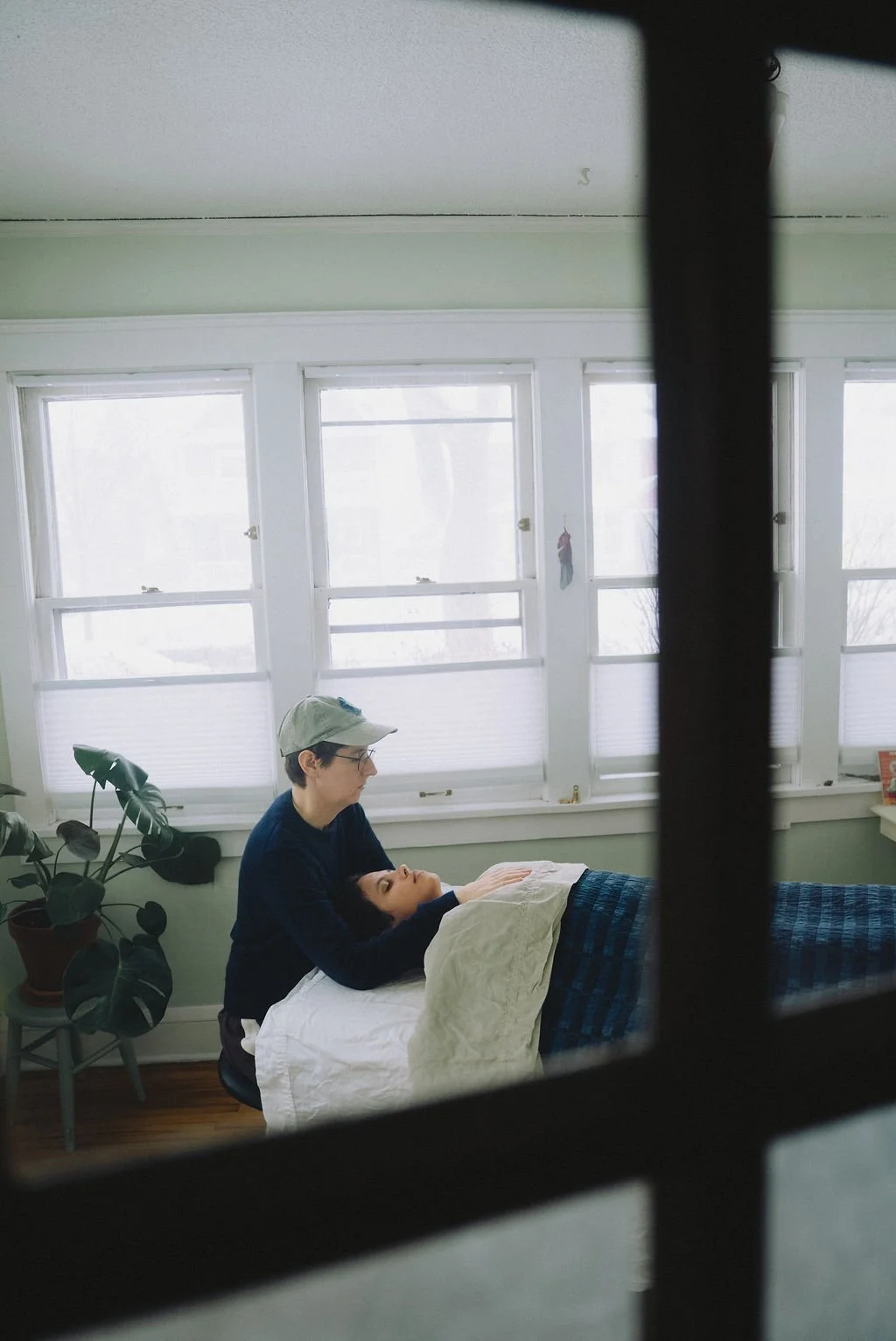 A person wearing glasses and a hat leans over another person lying in bed, in a room with multiple windows and a potted plant.