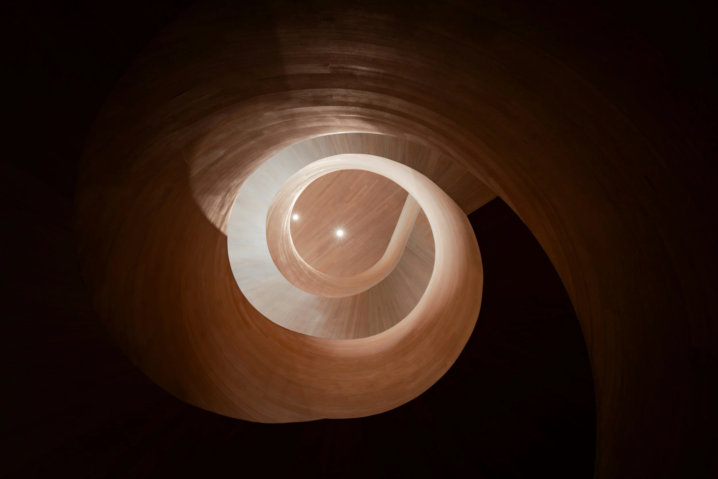 An upward view of a wooden spiral staircase with a circular opening at the center, illuminated by small lights.