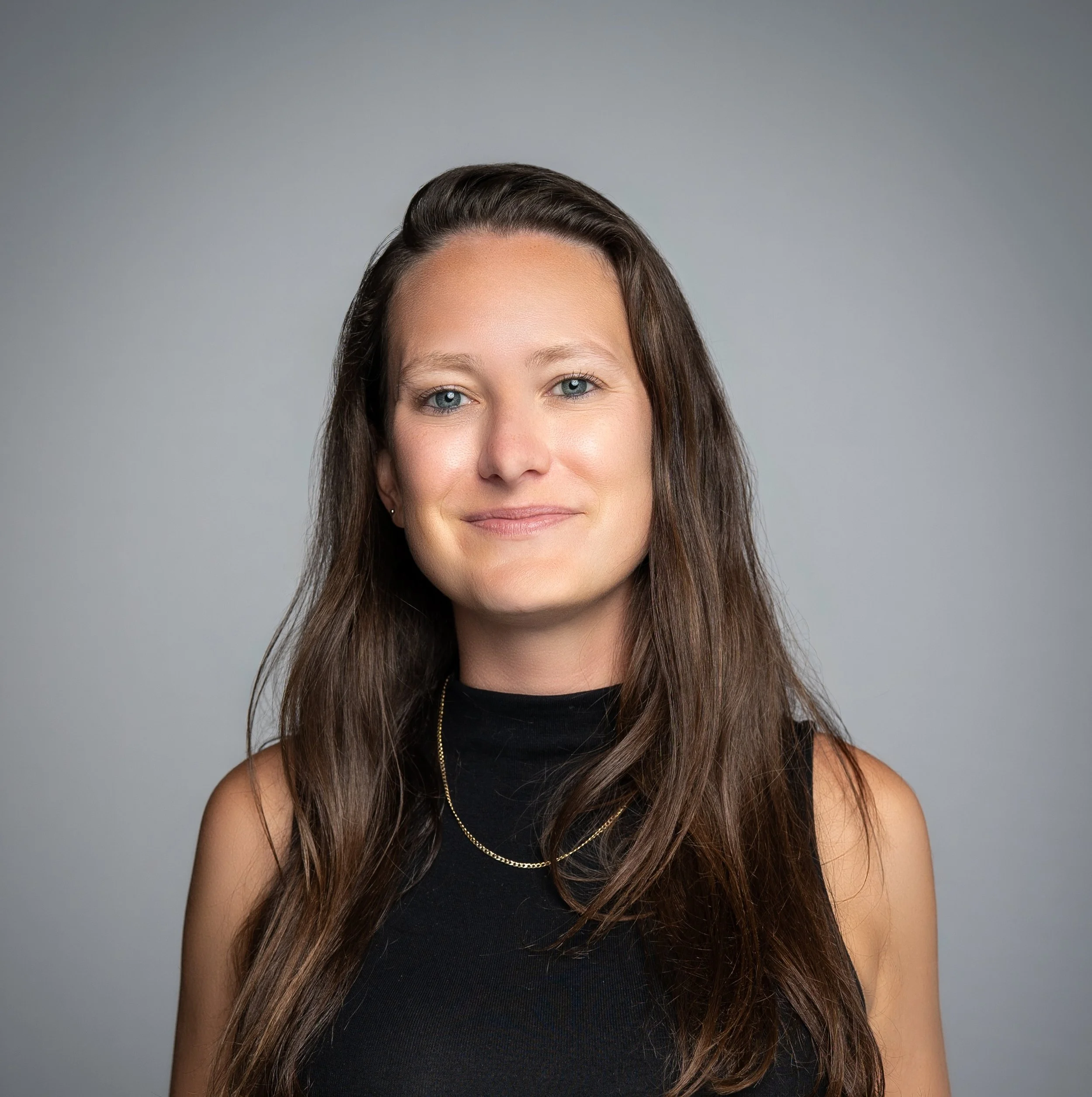 A woman with long brown hair, light skin, and blue eyes wearing a black sleeveless top and layered gold necklaces, smiling softly against a gray background.