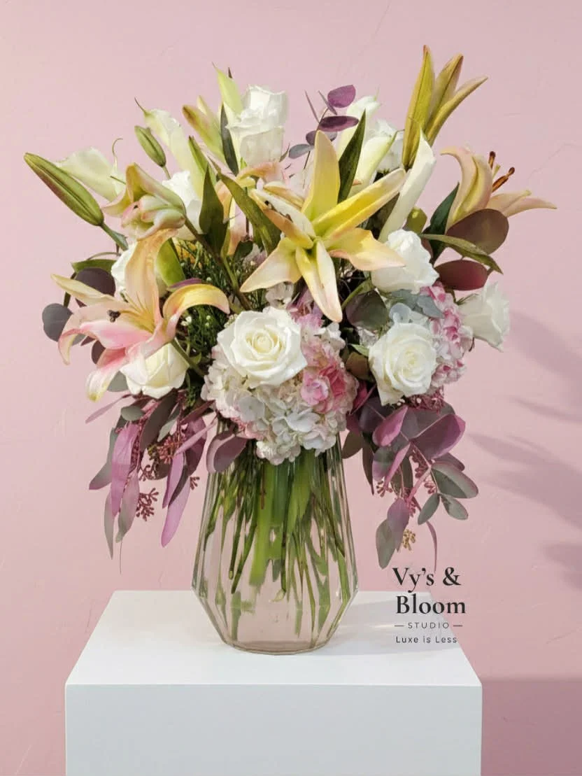 A floral arrangement in a transparent vase with white roses, pink hydrangeas, light pink lilies, and various greenery, placed on a white pedestal against a pink wall.