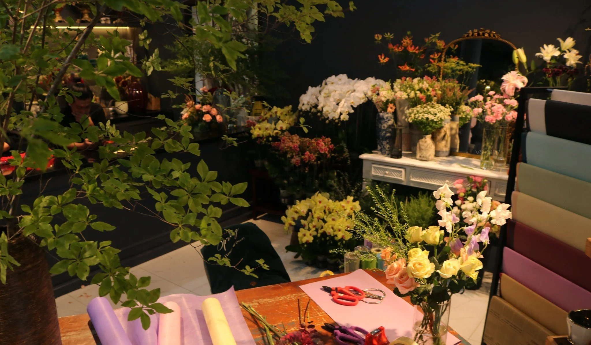 Interior of a flower shop with colorful flowers, floral arrangements, wrapping paper, and gardening tools on a wooden table, surrounded by various floral displays and supplies.