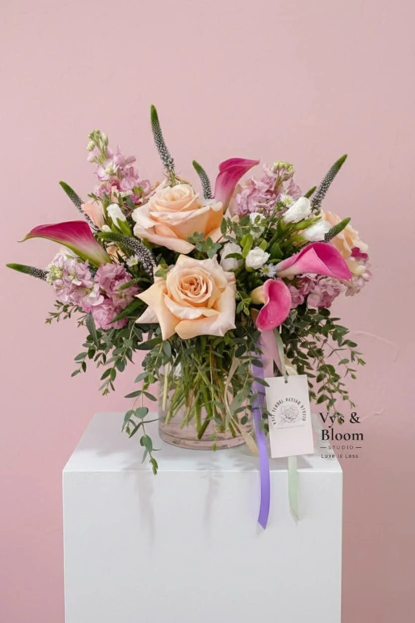 A pink and peach floral arrangement with roses, calla lilies, and greenery in a glass vase, placed on a white pedestal against a pink wall.