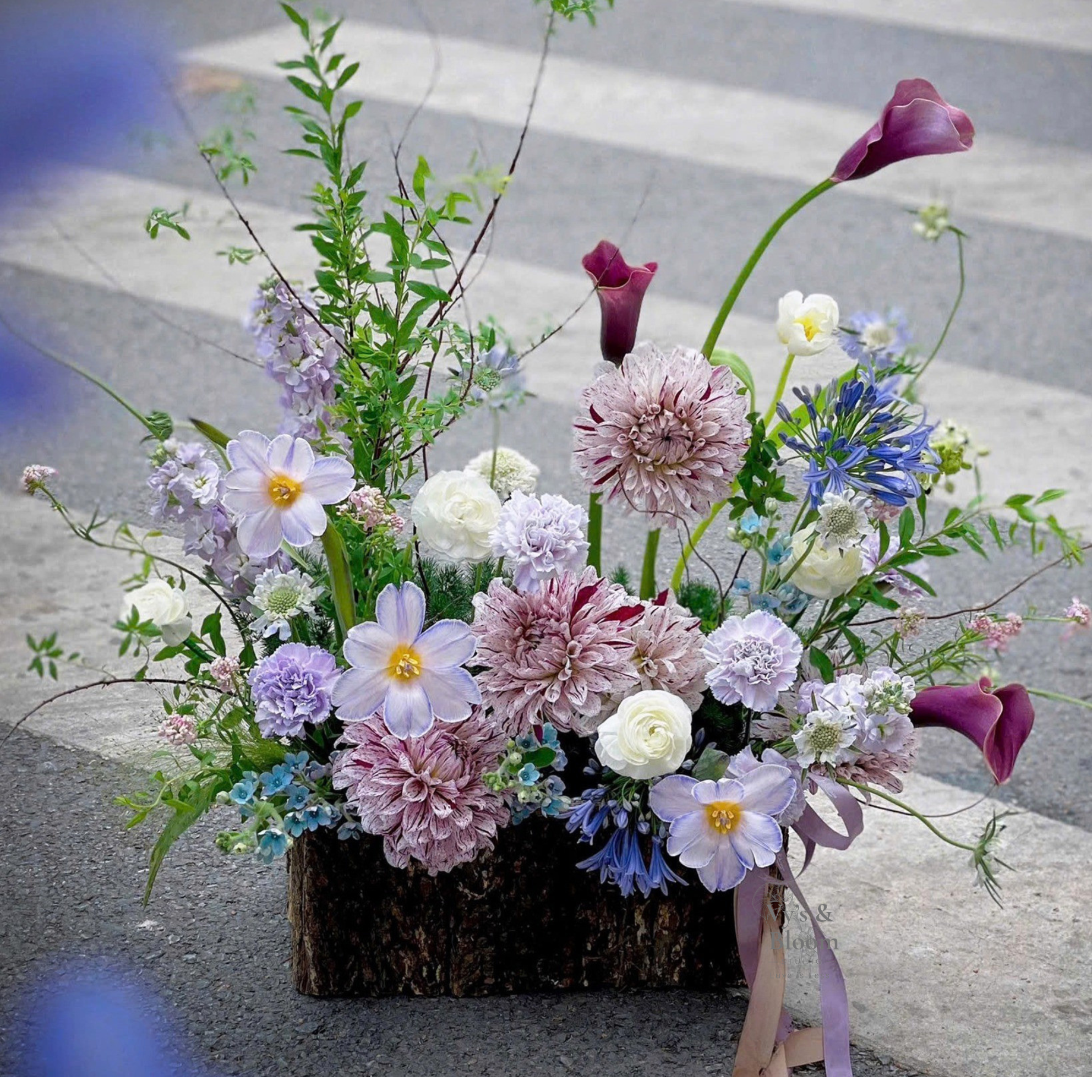 Bouquet of mixed flowers including calla lilies, dahlias, forget-me-nots, and other blooms in a wooden box on a stone surface.