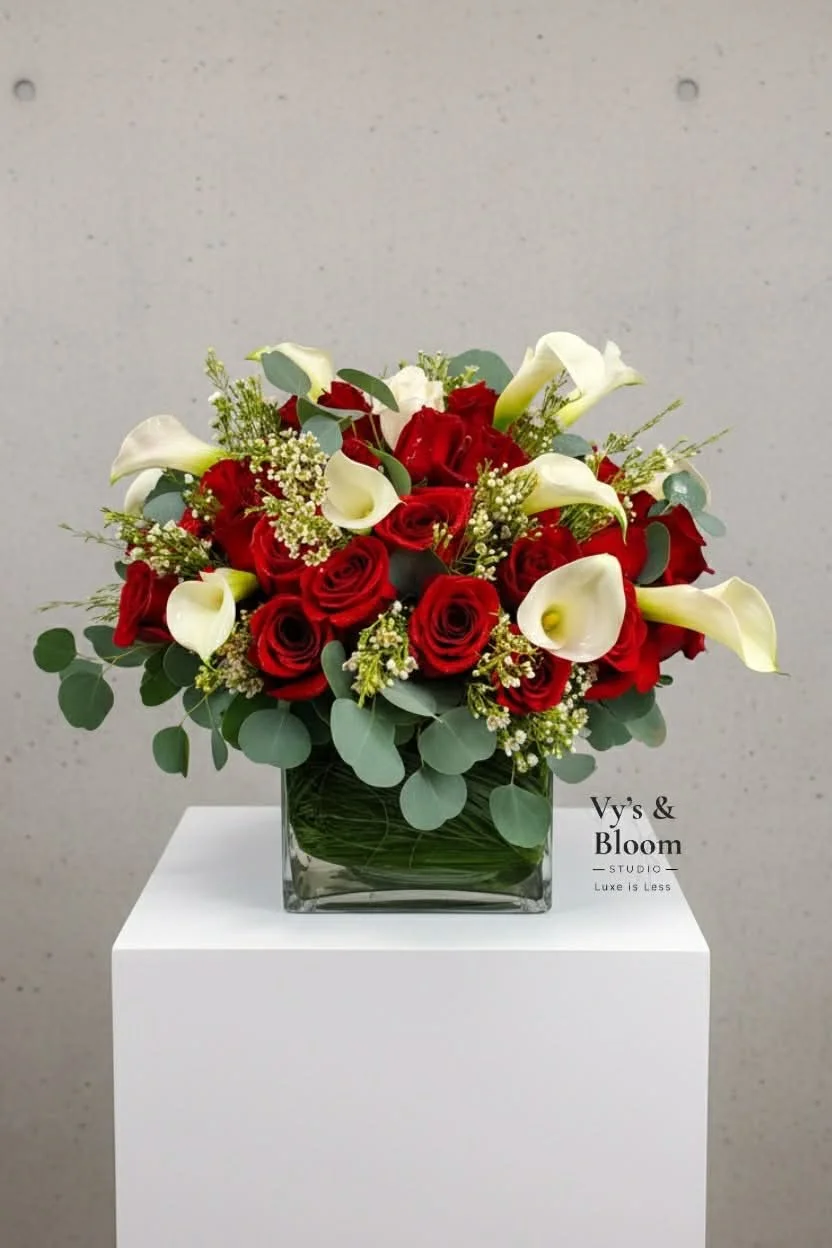 A floral arrangement featuring red roses and white calla lilies in a clear glass square vase, accented with eucalyptus leaves, displayed on a white pedestal.