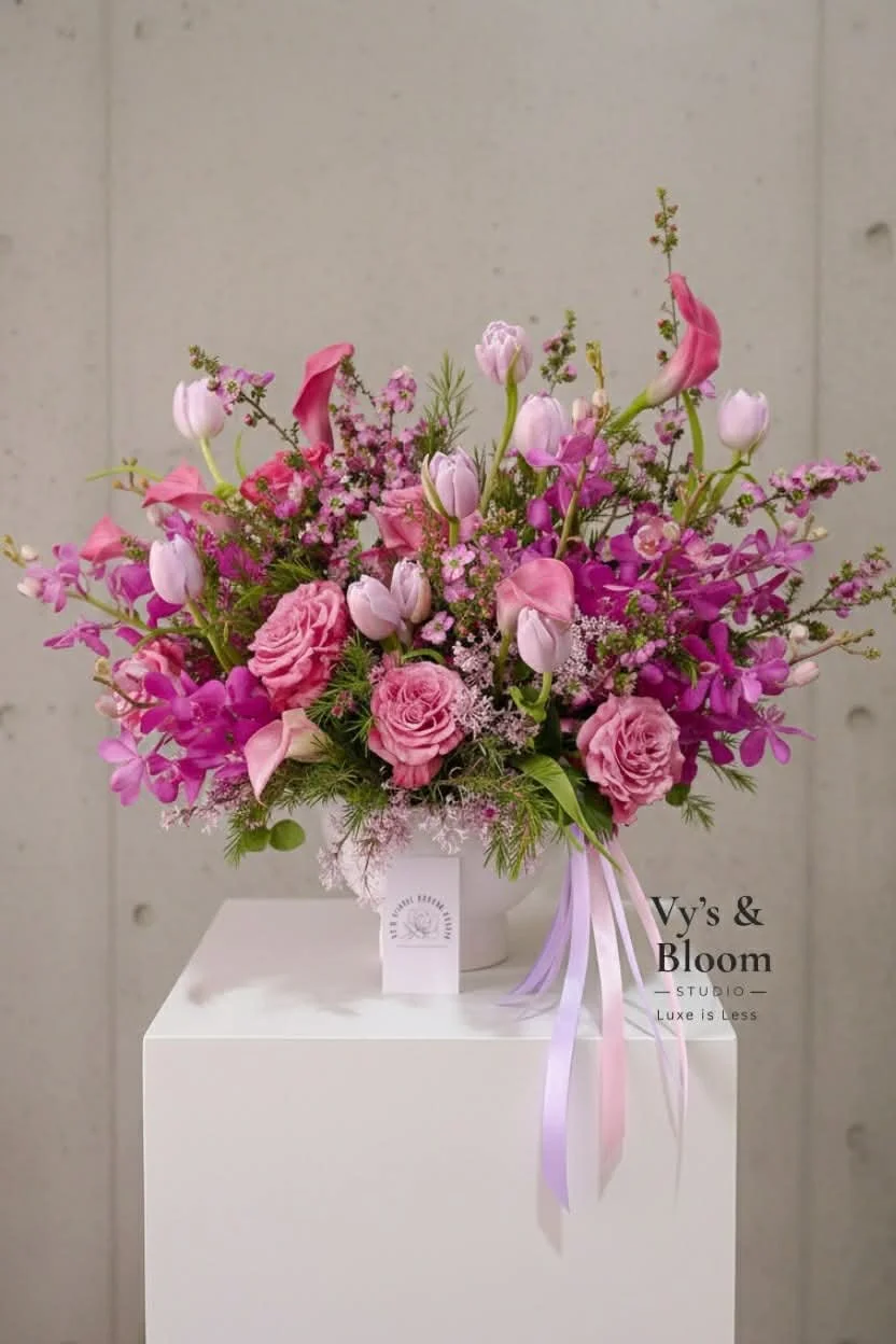 Pink and purple flower bouquet in a white vase on a white pedestal, with a gray wall background.