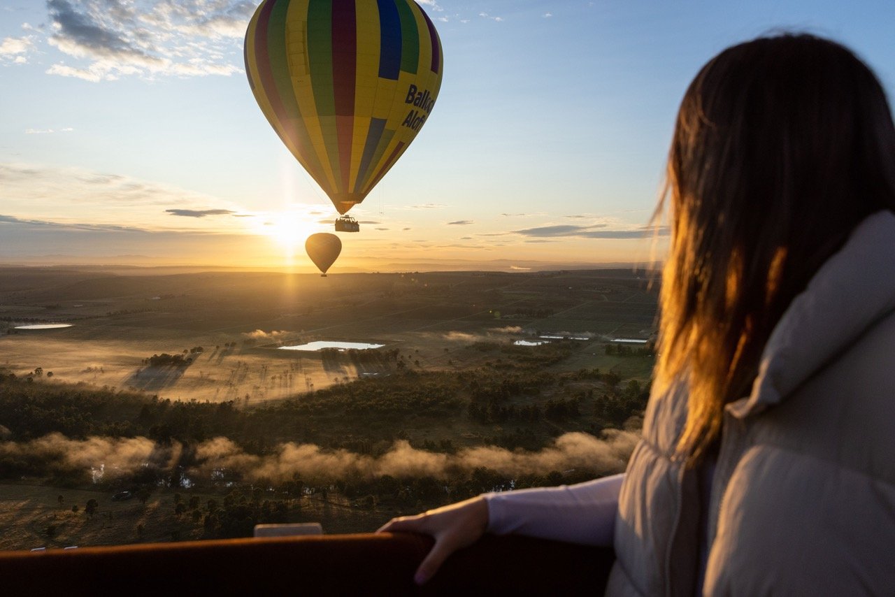 A person watching hot air balloons in the sky at sunrise over a landscape with fields and lakes.