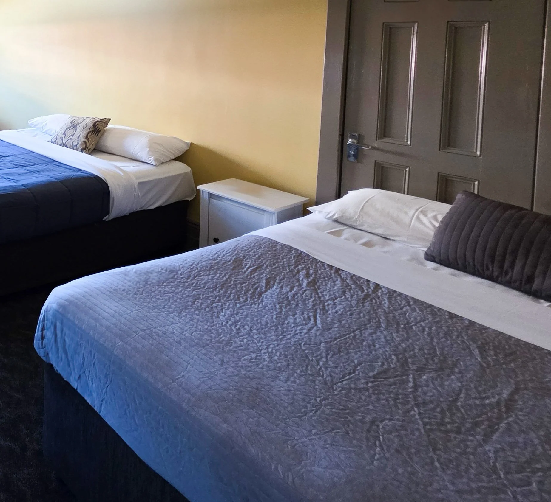 Two beds with white and grey bedding, a nightstand, and pillows against a yellow wall in a bedroom. A closed brown door is visible on the right side of the image.