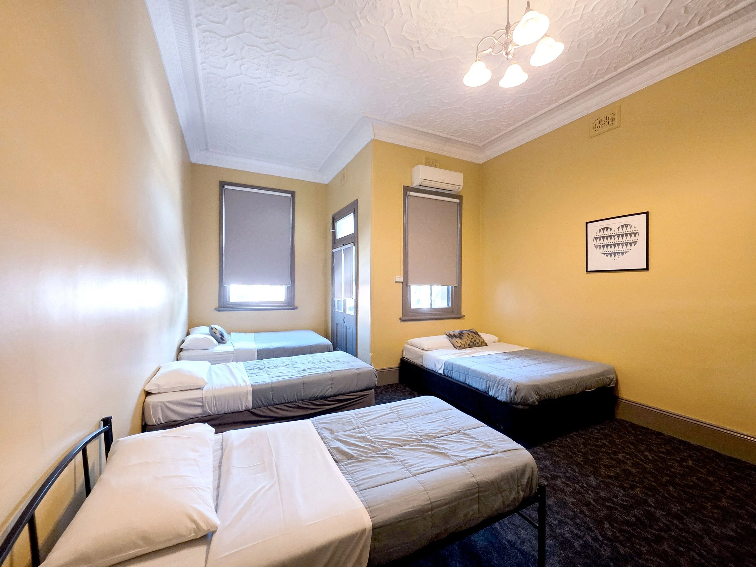 A room with yellow walls and ornate white ceiling molding, containing four beds with white sheets and pillows. There are two windows with gray frames and blinds, a framed art piece of a heart made of triangles, and a ceiling light fixture with multiple bulbs.
