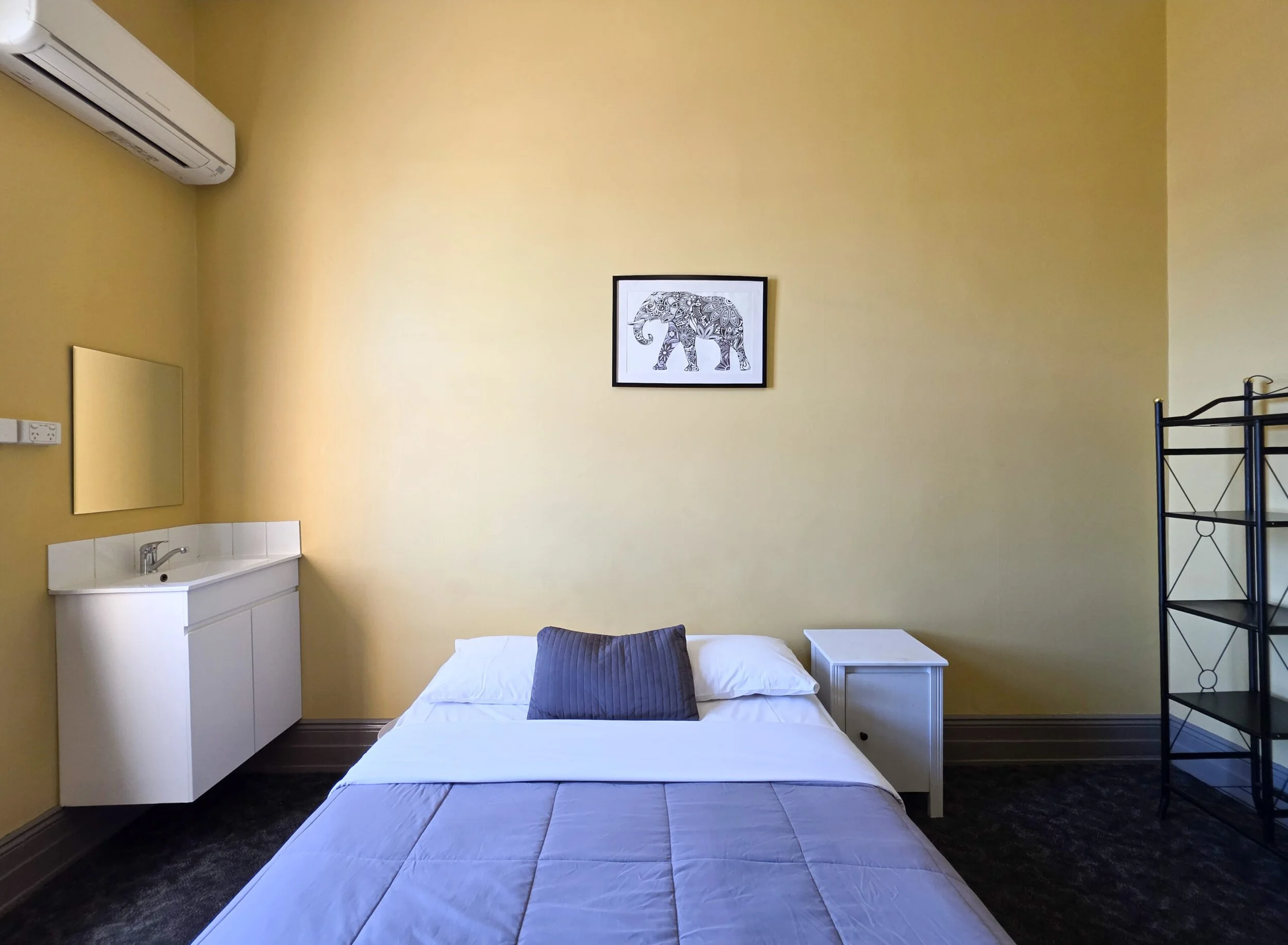 A small bedroom with yellow walls, a bed with white bedding and a purple pillow, a white nightstand, a black metal shelf, and framed artwork of an intricately designed elephant on the wall.