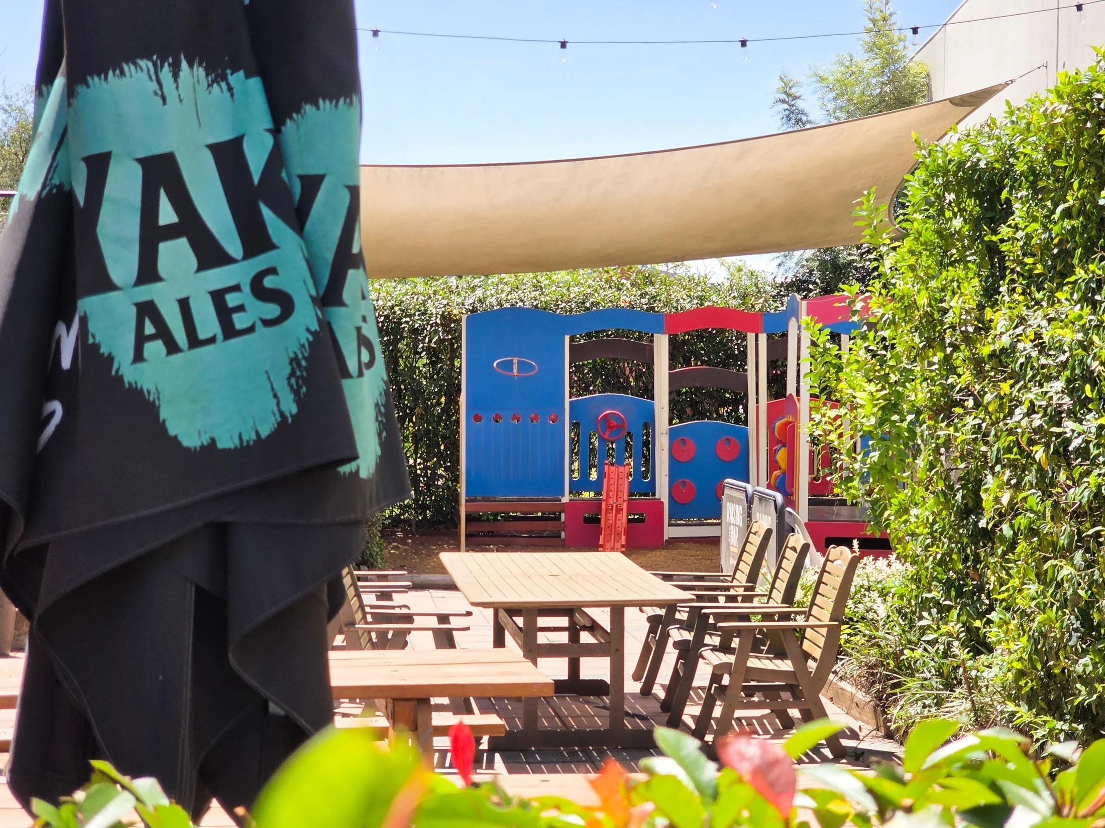 Part of a person wearing a black and turquoise shirt, standing outdoors in a patio area with tables and chairs, trees, bushes, a colorful play structure, and a shade sail.