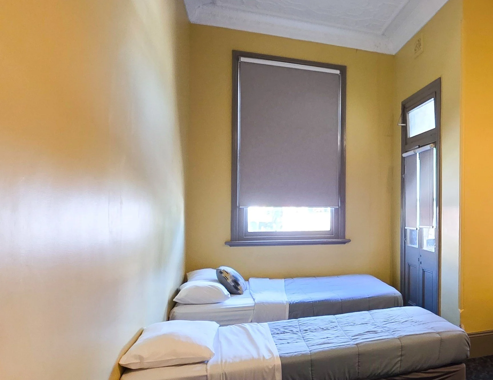 A bedroom with yellow walls, a window with a roller shade, a door with a screen, a single bed with white sheets and pillows, and a decorative pillow with a face print.