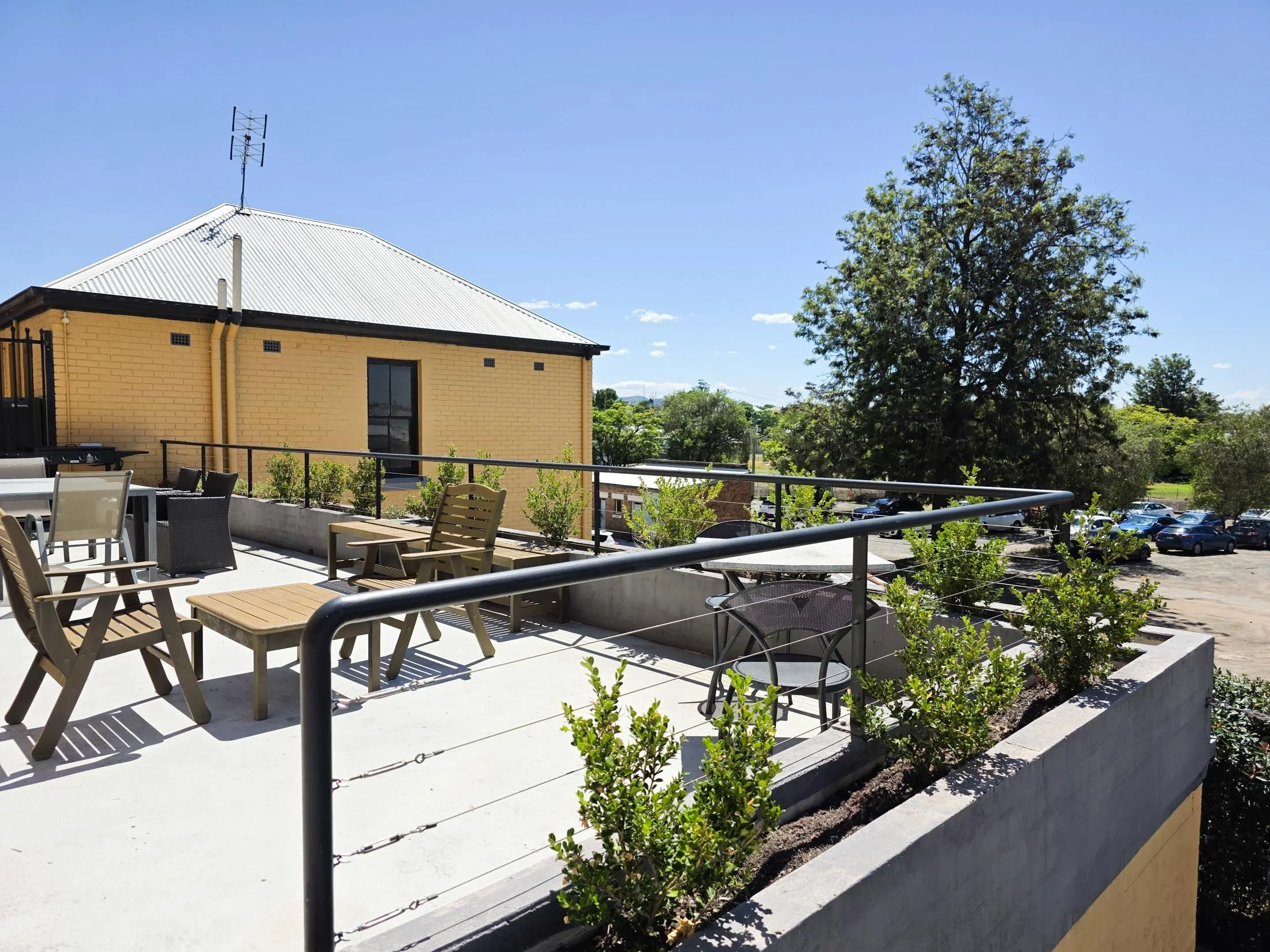 Outdoor patio area with chairs and tables, surrounded by small plants and a low wall, with a yellow brick building and a large tree in the background under a clear blue sky.