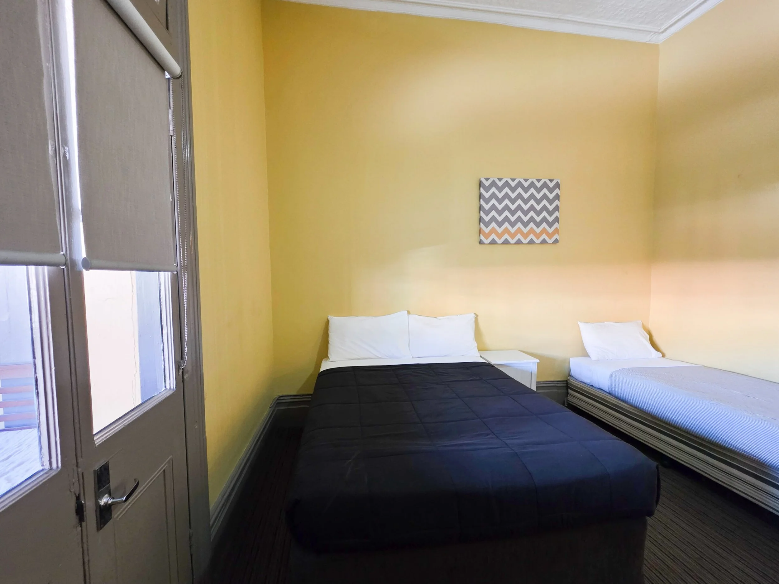 A bedroom with yellow walls, two beds with white pillows, one with a black quilt, and the other with a striped bedspread. There is a small white nightstand next to the beds and a piece of artwork with a geometric pattern hanging on the wall. The room has windows with blinds and a door with a mail slot and lock.