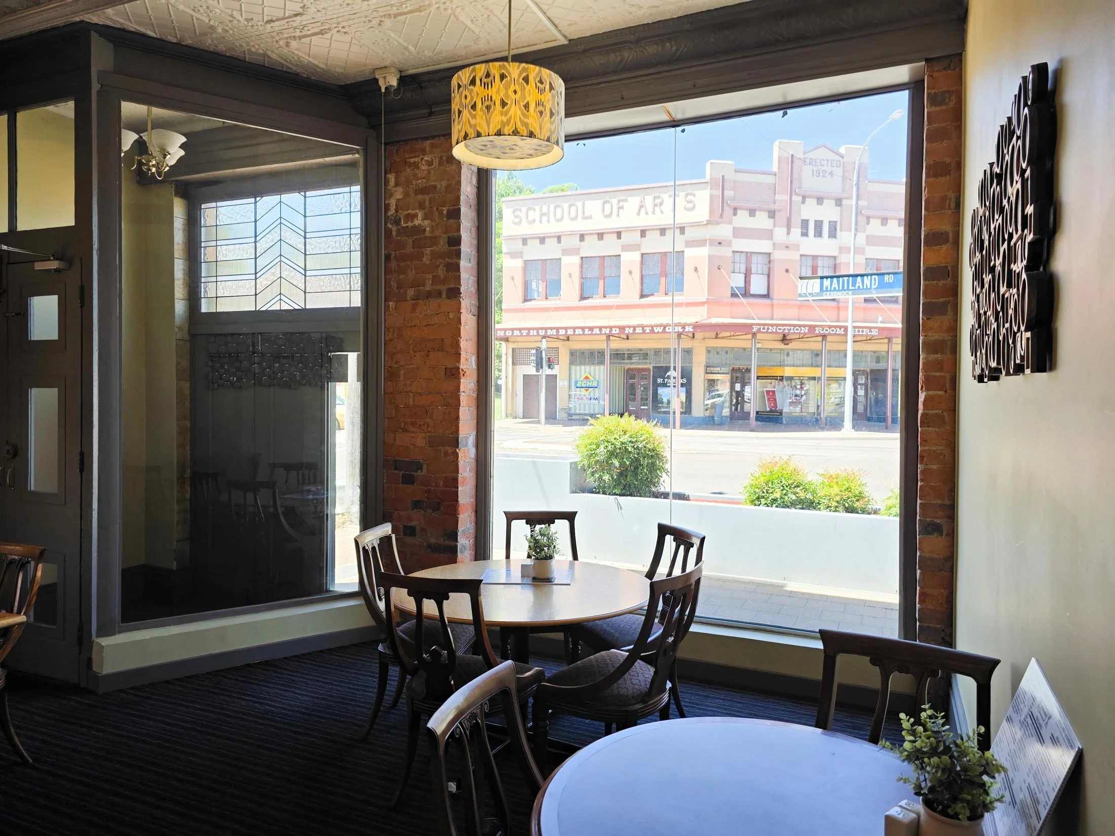 Interior of a cafe or restaurant with round tables, chairs, and large windows showing a street view including a building labeled 'School of Arts' and street signs.