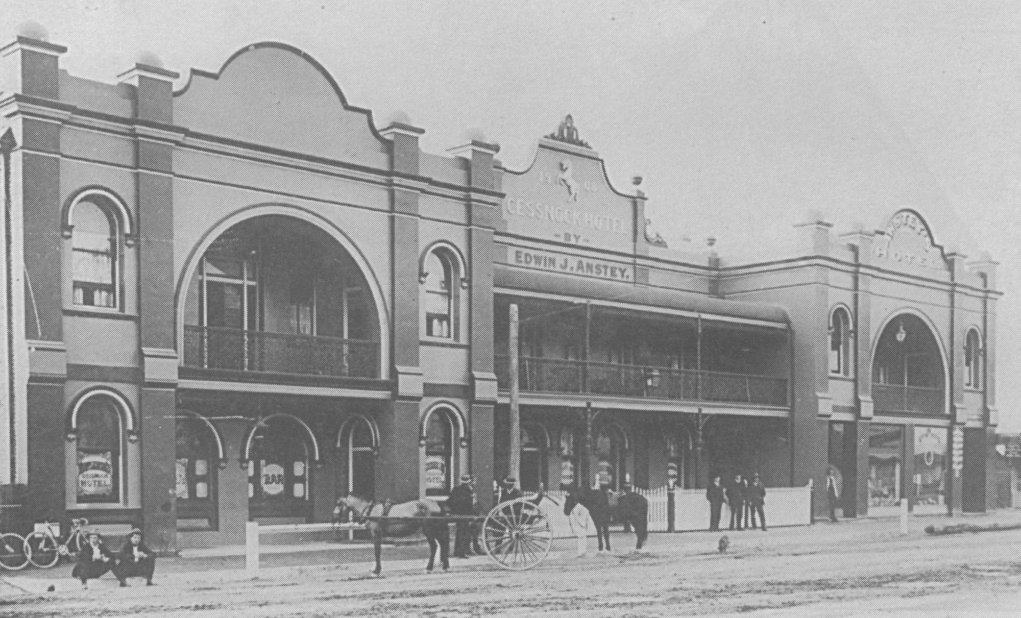 Historical black and white photograph of a two-story building with arched windows and a sign that reads "Edwin G. Austry." In front, there are horses, a horse-drawn carriage, bicycles, people, and a fence. The building appears to be from the early 20th century.