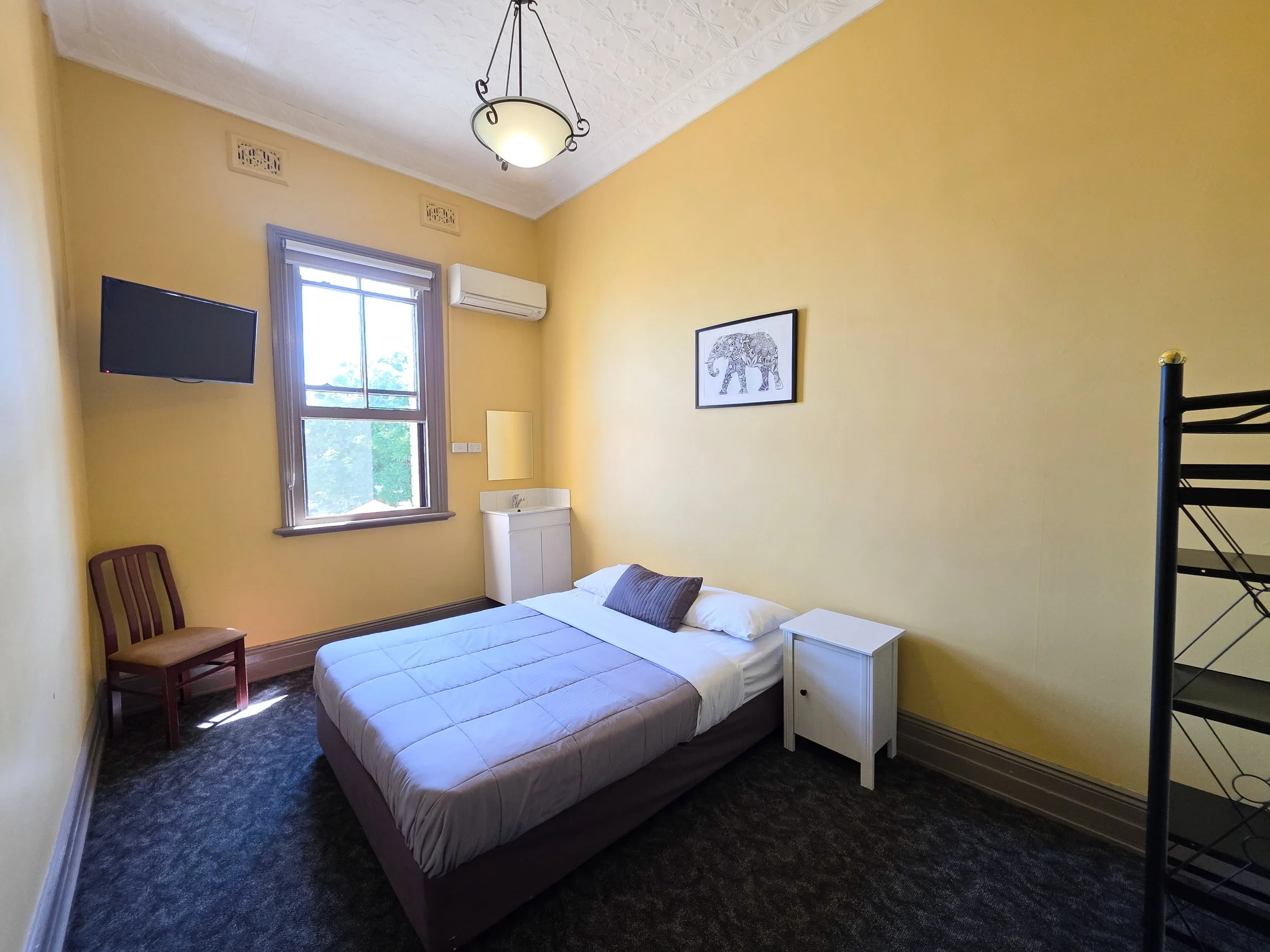 A small bedroom with yellow walls, a window, a ceiling light, a wall-mounted TV, a bed with white bedding and dark pillows, a small white nightstand, a framed picture of an elephant, a wooden chair, and a black metal staircase.