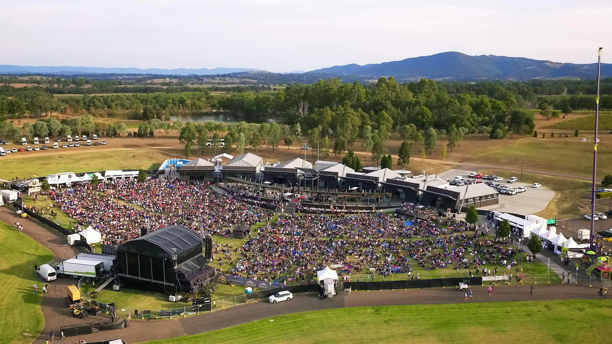 An outdoor concert or event with a large crowd seated on the grass near a stage and various tents, set in a scenic area with trees, water, and mountains in the background.