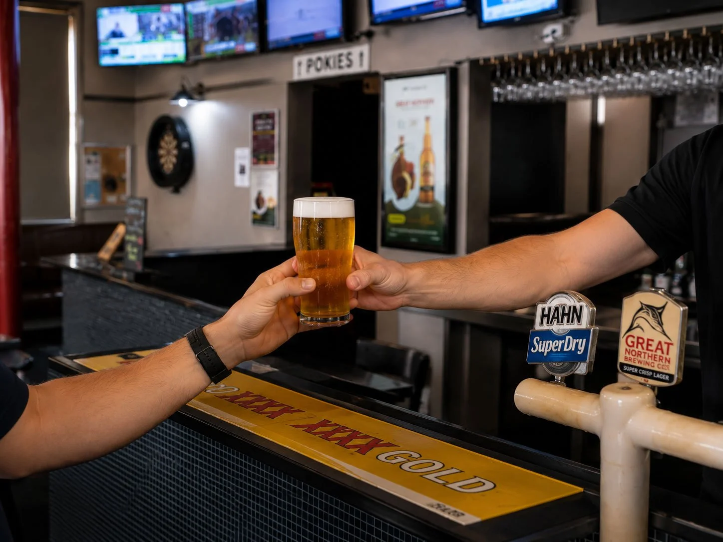 A person receiving a glass of beer from a bartender at a bar counter. The bar has assorted taps and TV screens displaying sports in the background.