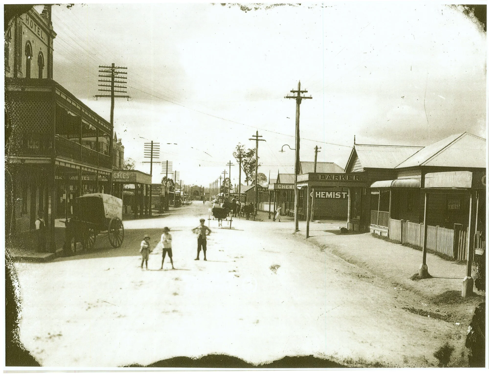 A black and white vintage photograph of a street scene from the early 20th century. It features several children playing in the street, horse-drawn wagons, and storefronts including a chemist shop. The street is lined with wooden buildings and utility poles, with a prominent two-story building on the left side.