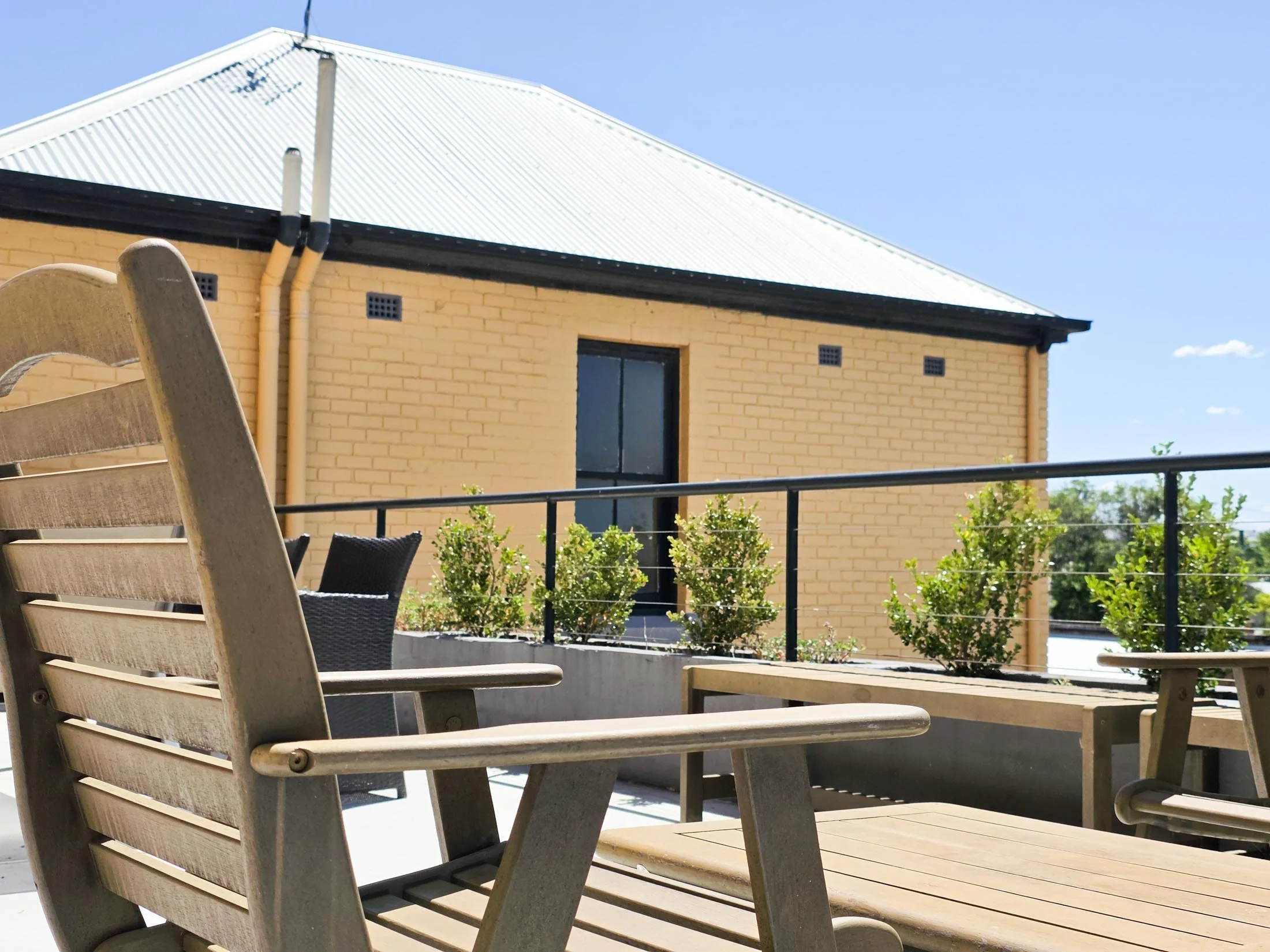 Patio furniture with a wooden chair and a table in front of a house with a yellow brick wall and a metal roof, under a clear blue sky.
