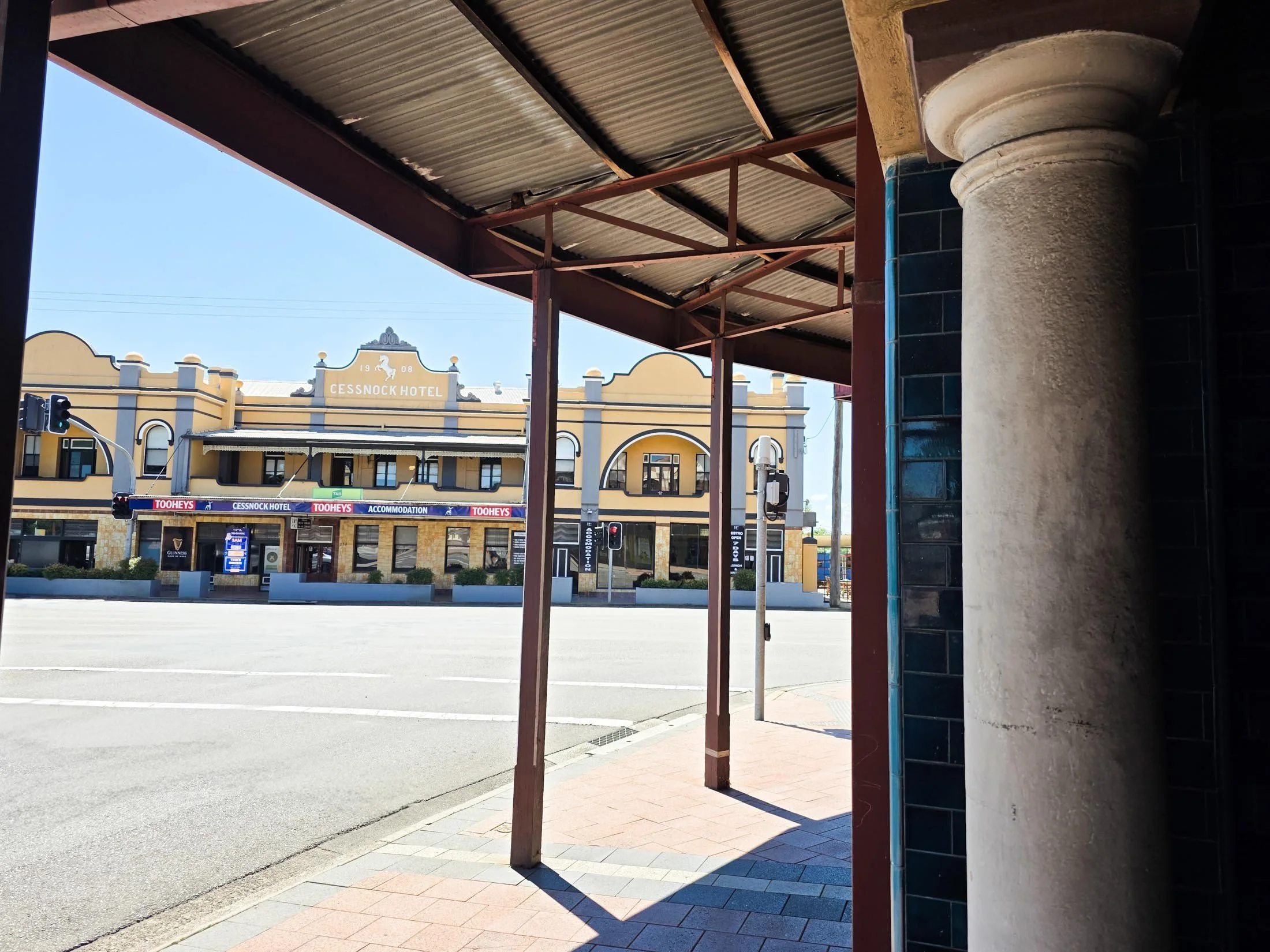 View from a shaded sidewalk looking into a street with a building named Cessnock Hotel across the road, featuring a yellow facade and decorative architectural details.