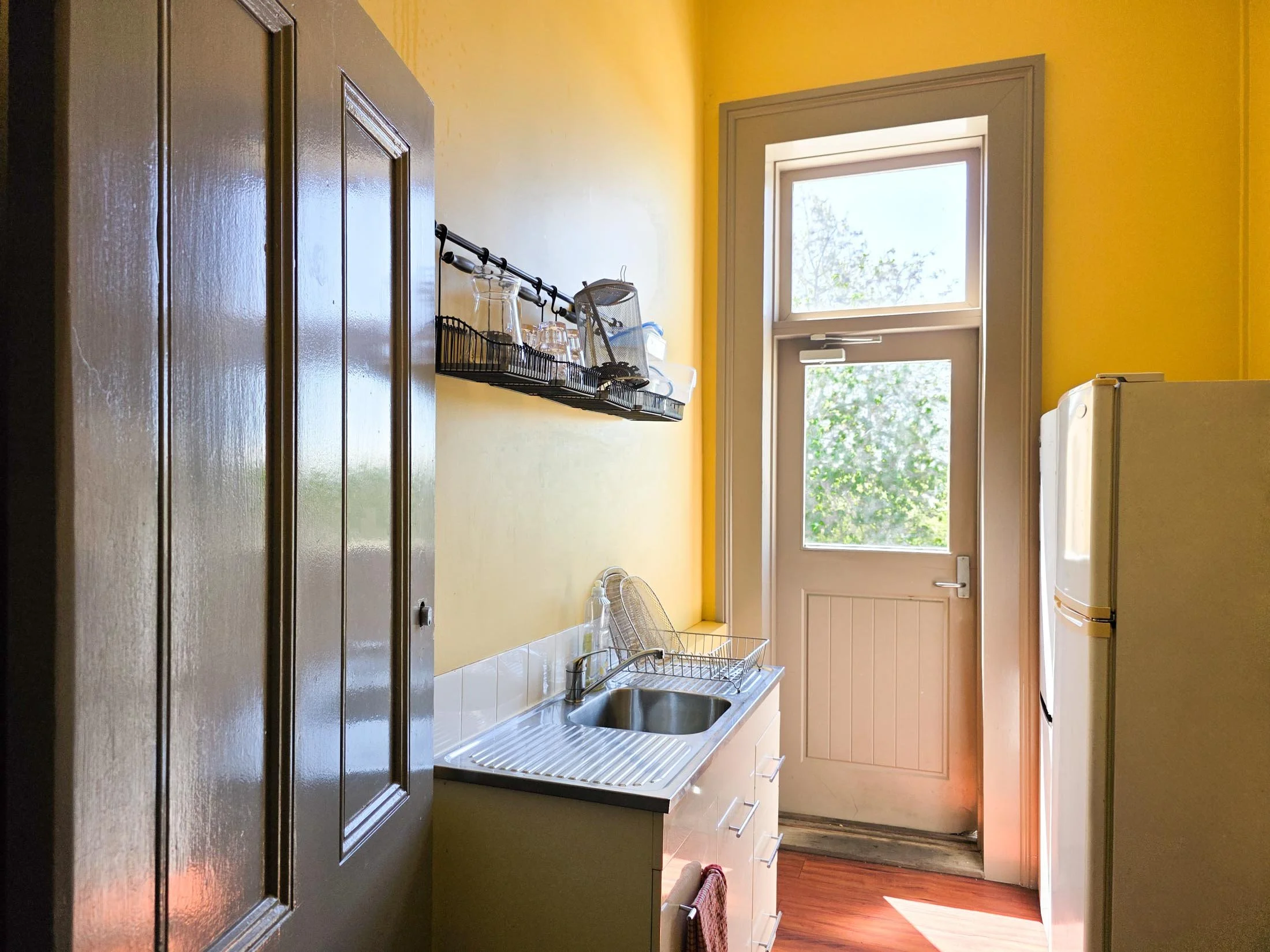 A small kitchen with bright yellow walls, a white sink with a drying rack, a window with a view of green trees, a countertop with dishes, and a fridge on the right side.