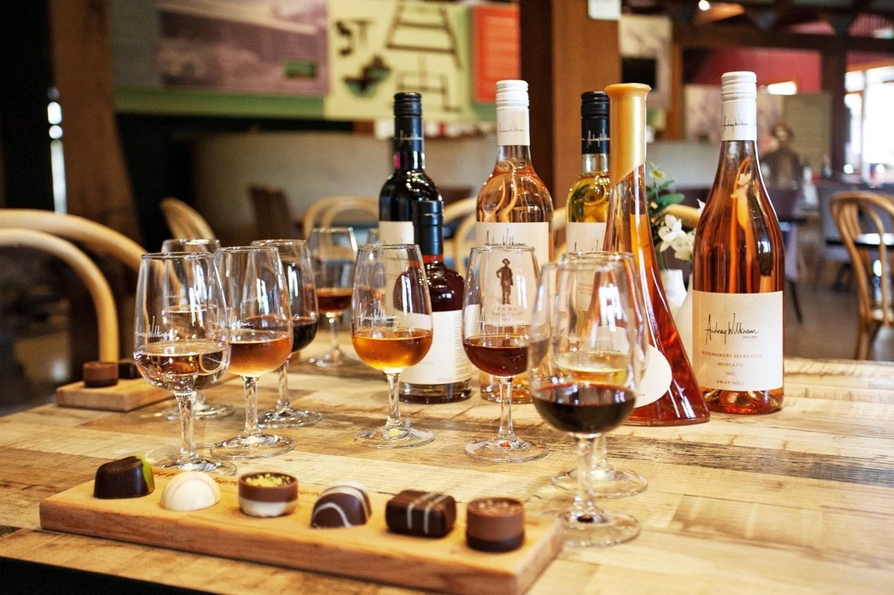 A wooden table set with various bottles of wine and multiple wine glasses filled with rosé, red, and white wine. There is a tasting tray of assorted chocolates in the foreground and a blurred background of a cozy restaurant or wine bar interior.