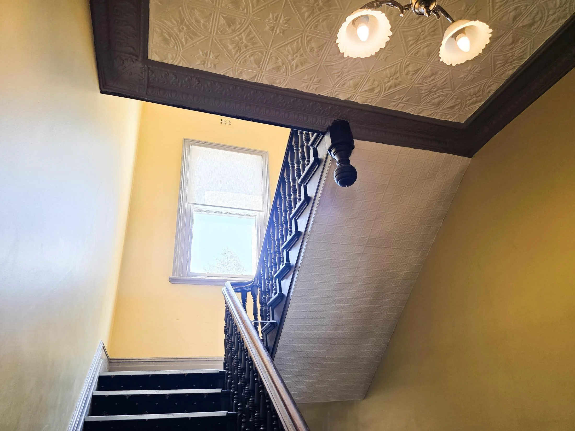 Interior view of a staircase in a house with yellow walls, a window at the top, and an ornate ceiling with a chandelier.