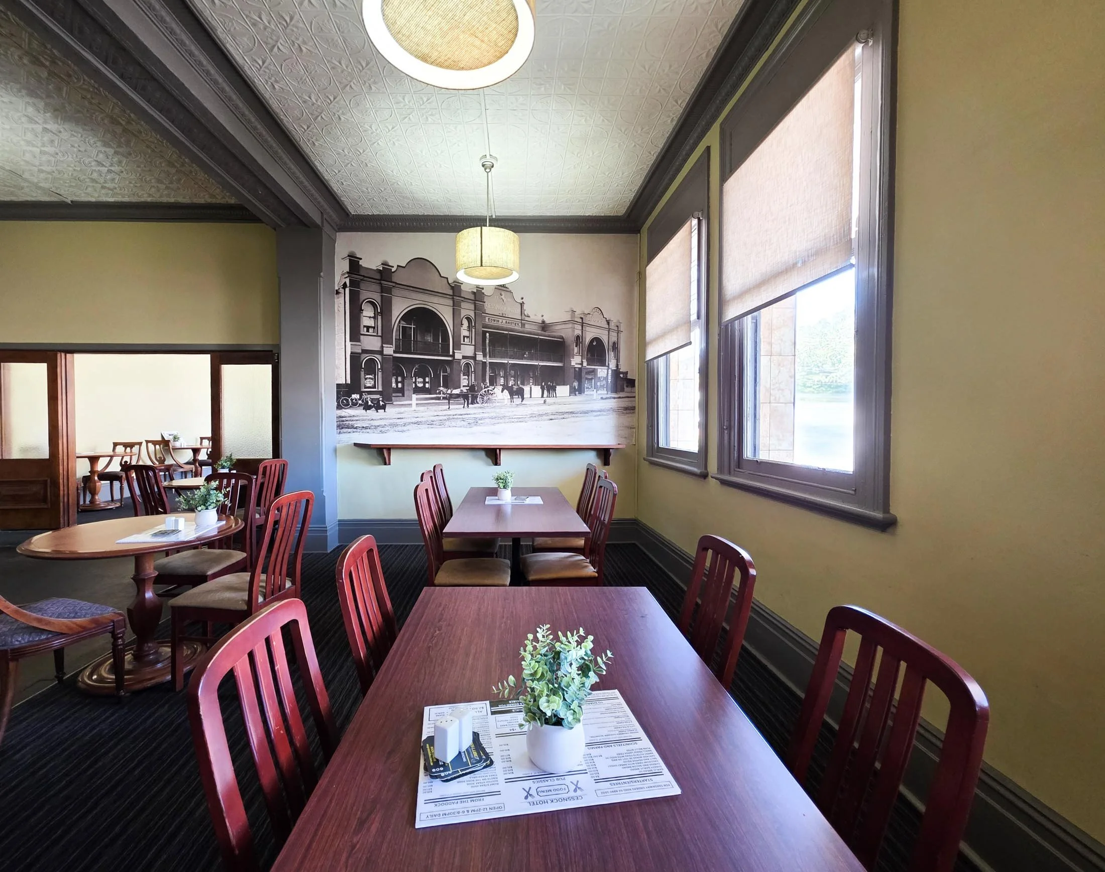 Interior of a restaurant with wooden tables, red chairs, potted plants, and a wall clock. Bright windows with blinds; a vintage mural of a historic street scene on the wall.