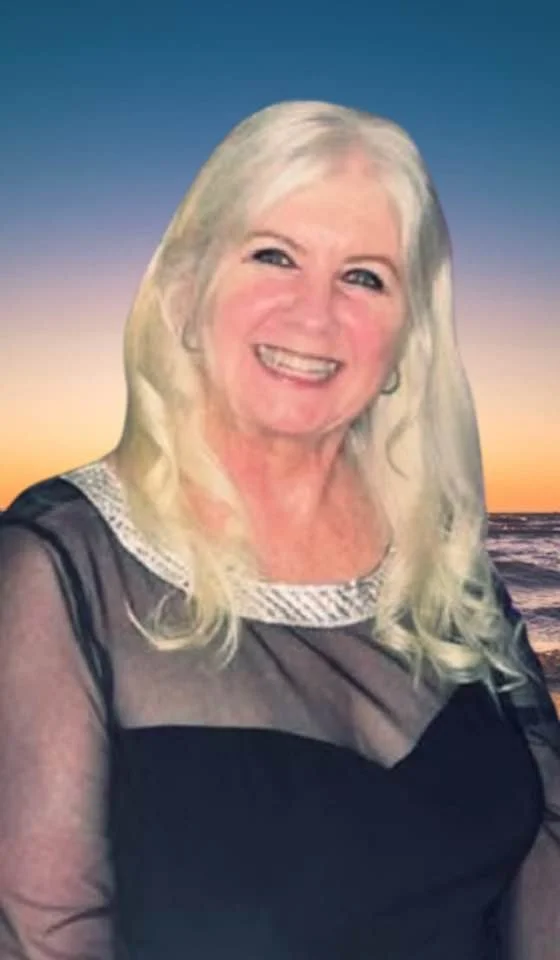 A smiling woman with long blonde hair, wearing a black sheer top with a decorative collar, standing outdoors at sunset with a beach and ocean in the background.