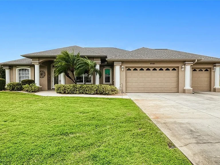 Single-story suburban house with beige exterior, three-car garage, and a well-maintained front yard with green grass, bushes, and a palm tree, under a clear blue sky.