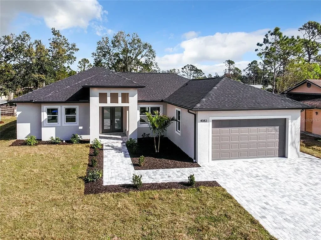 Modern single-story house with white exterior walls, dark gray shingle roof, front yard with small plants, and a paved walkway leading to the front door.