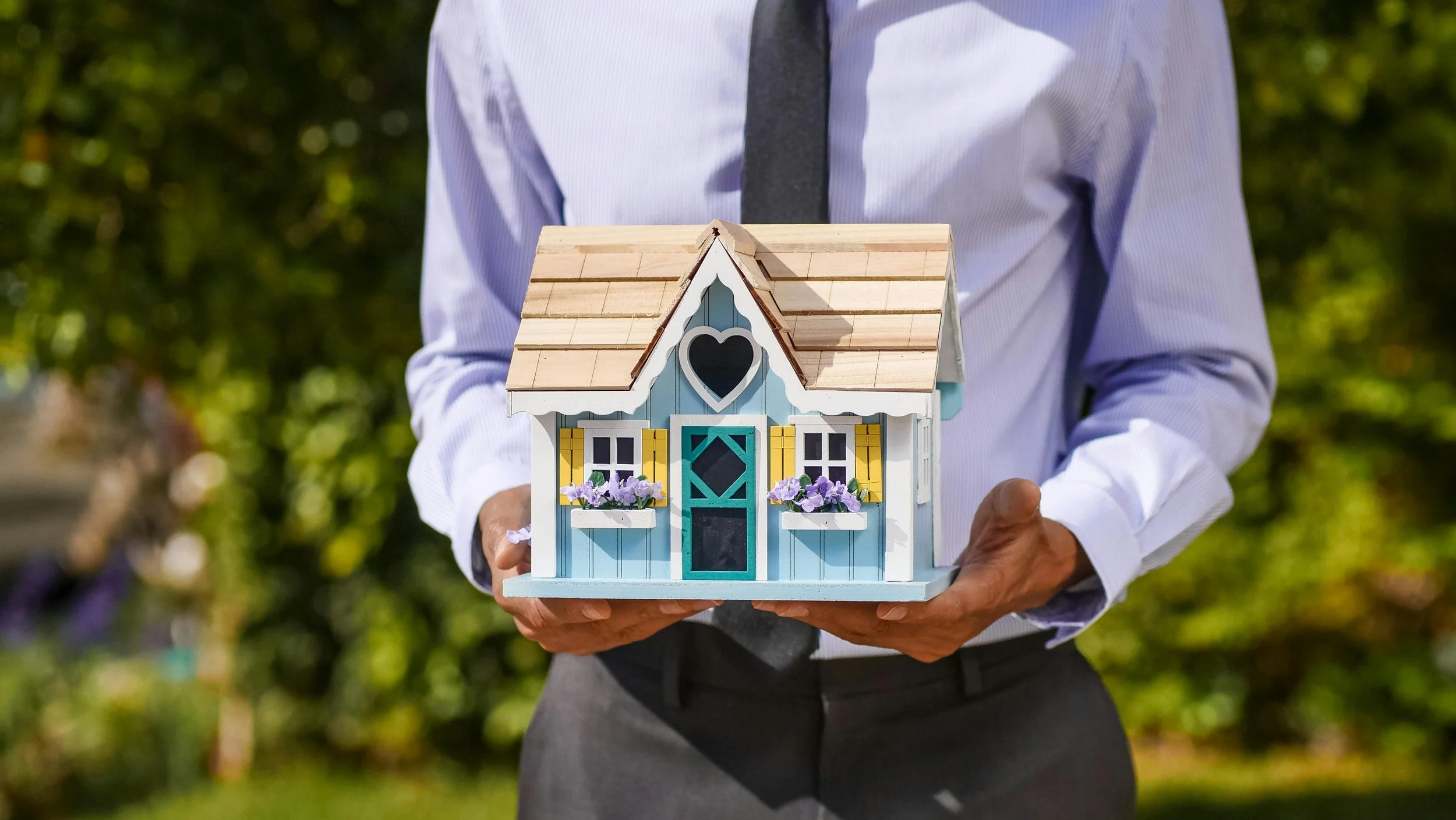 Person holding a colorful miniature house with a heart-shaped window, yellow shutters, and purple flower boxes, outdoors on a sunny day.