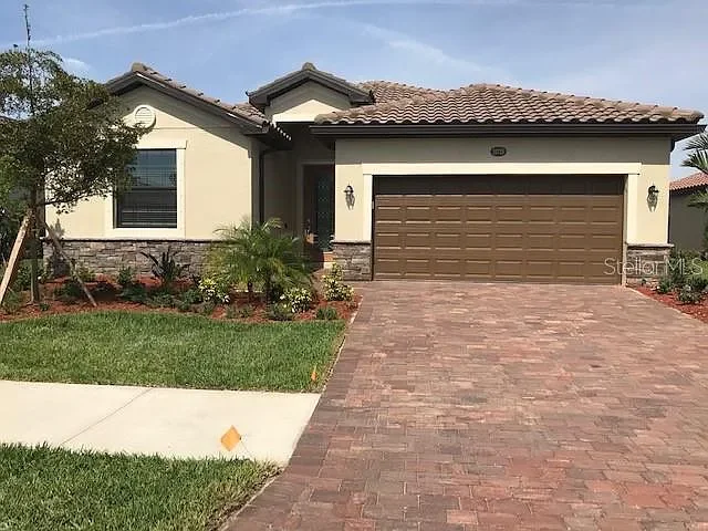 Front view of a single-story house with a brick driveway, a two-car garage, and a small landscaped yard with grass and plants.